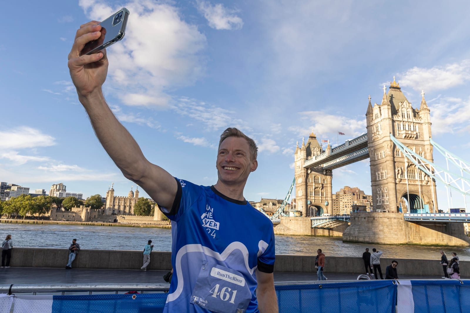 A smiling runner wearing a blue race shirt takes a selfie by the Thames River with London's Tower Bridge in the background on a sunny day.