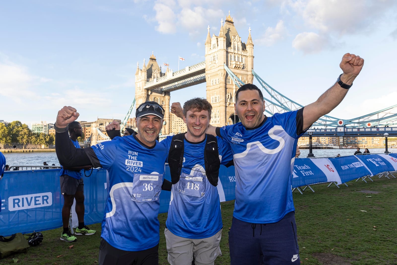 Three smiling runners in blue "Run the River 2024" shirts celebrate with raised arms in front of Tower Bridge in London, with a bright sky and race banners in the background.