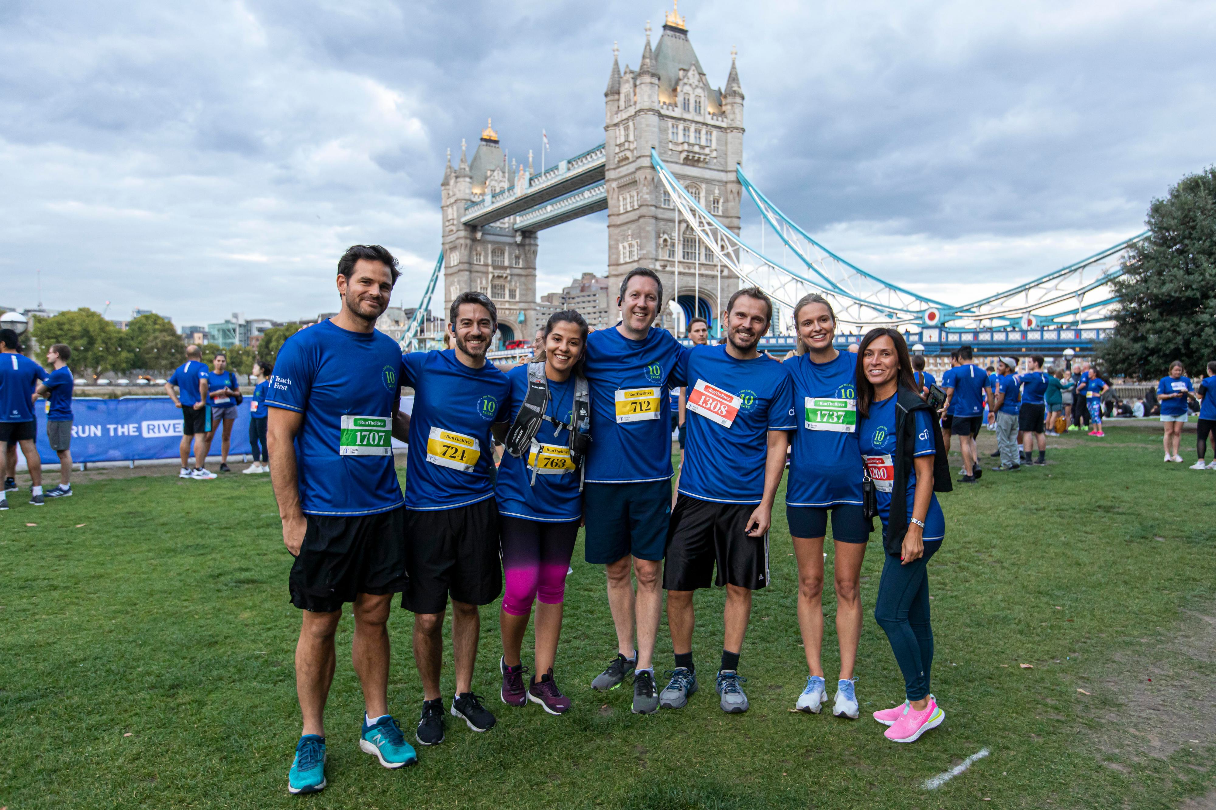 A group of seven people dressed in matching blue athletic wear stands smiling in front of Tower Bridge. They all wear race bibs and pose together on a grassy area, suggesting participation in a running event. Other participants and the Thames River are visible in the background.