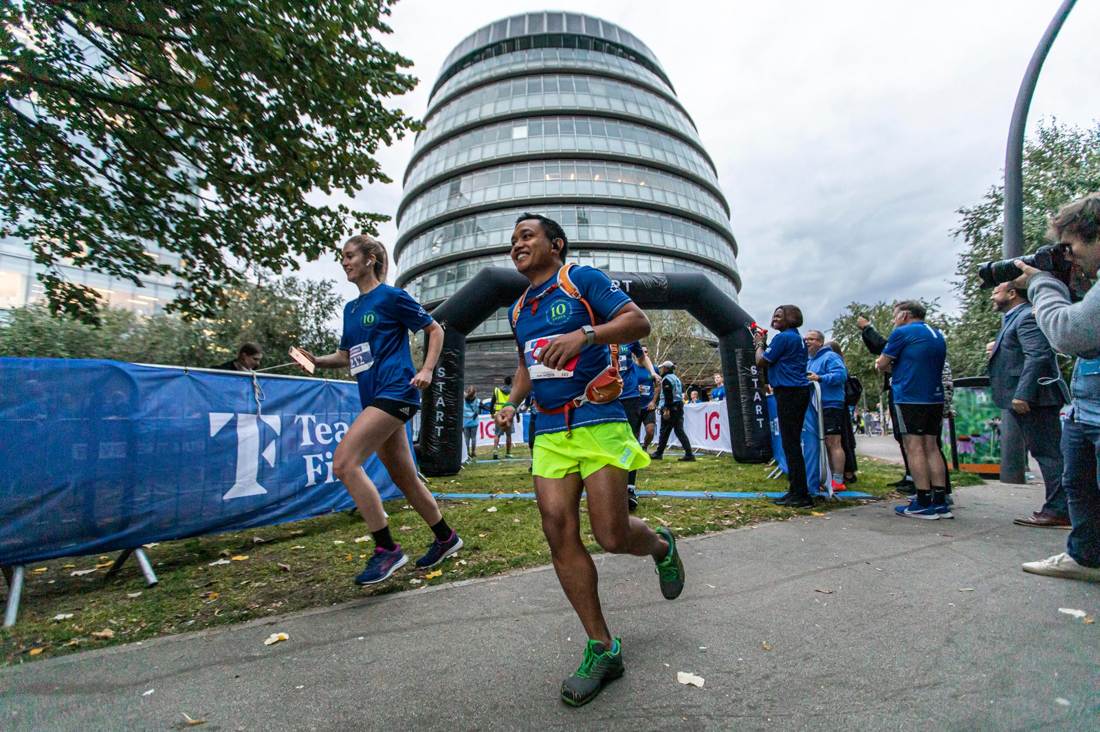 Two runners, wearing colorful athletic gear, participate in a race near a modern, rounded building. A photographer captures the moment nearby. Other participants and onlookers are visible in the background, and trees line the path.
