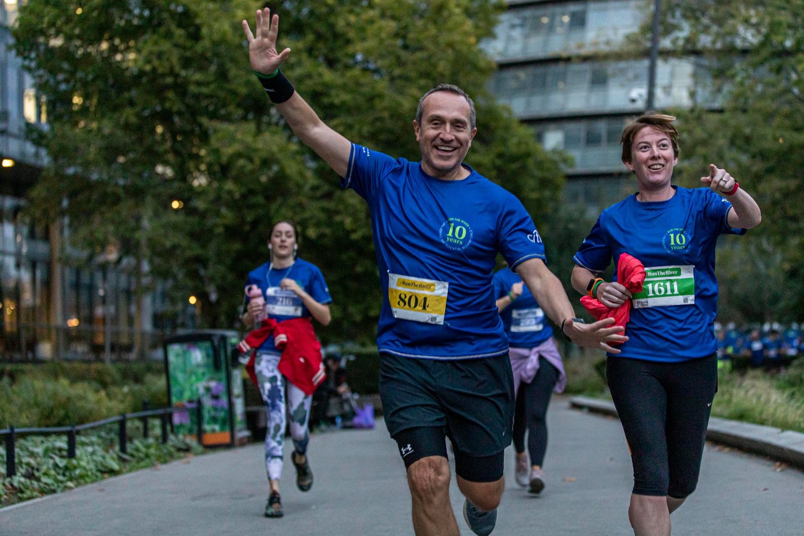 A group of runners is participating in a race. They are wearing blue shirts with numbers, running on a path through a park. A man in front, with a yellow bib number 804, is smiling and waving at the camera. Trees and buildings are visible in the background.