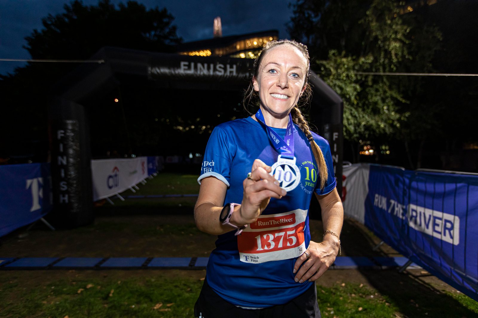 A runner in a blue jersey proudly displays her medal at the finish line of a race. She is wearing a race bib with the number 1375, and it is evening with a dark sky and lit trees in the background. She is smiling and appears to be content with her achievement.