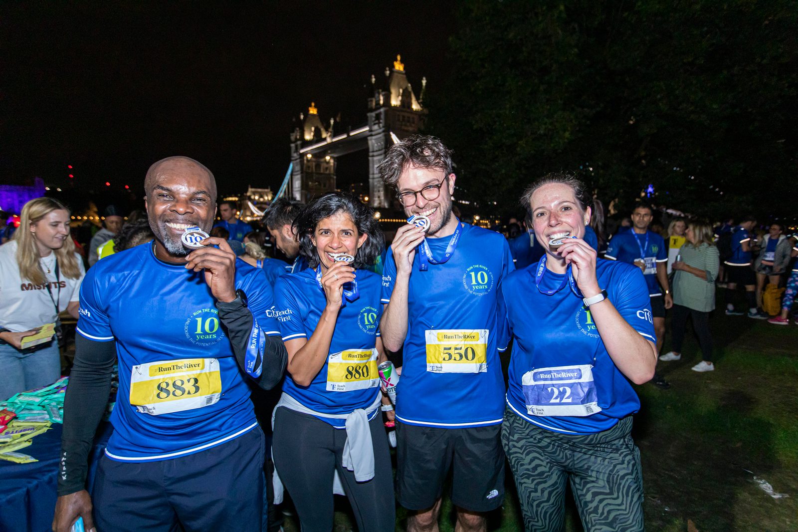 Four smiling runners wearing blue shirts, holding medals and posing for a photo at night in front of a landmark bridge and gathered crowd. They proudly display their medals by biting them. Their race bibs are visible, and other participants can be seen behind them.
