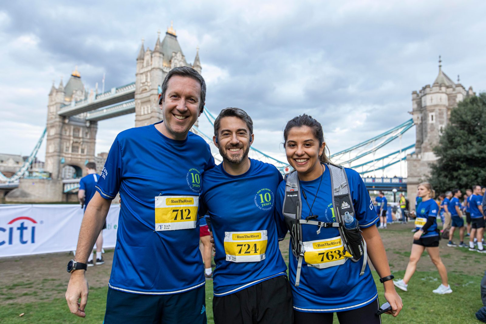 Three happy marathon runners wearing race bibs (numbers 712, 724, and 763) pose in front of Tower Bridge in London. They are wearing blue shirts and have medals around their necks. The sky is cloudy, and other runners are visible in the background.