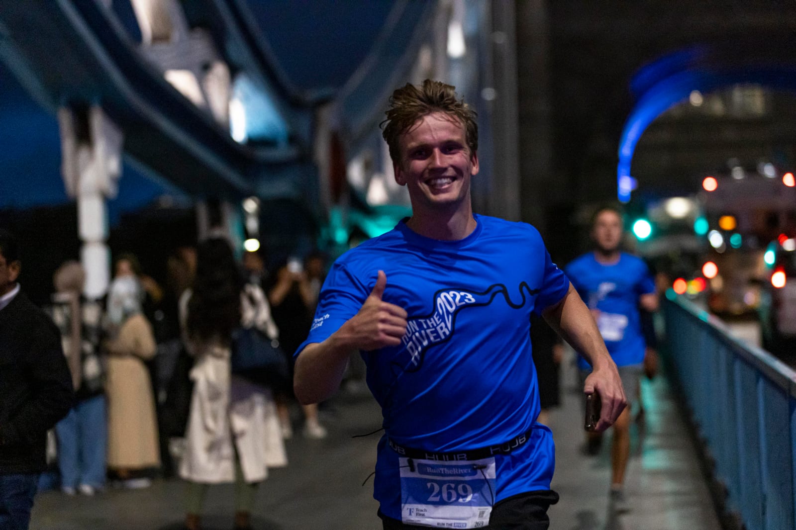 A man in a blue shirt and race bib smiles and gives a thumbs-up while running on a lit-up bridge at night, with other runners and spectators in the background.