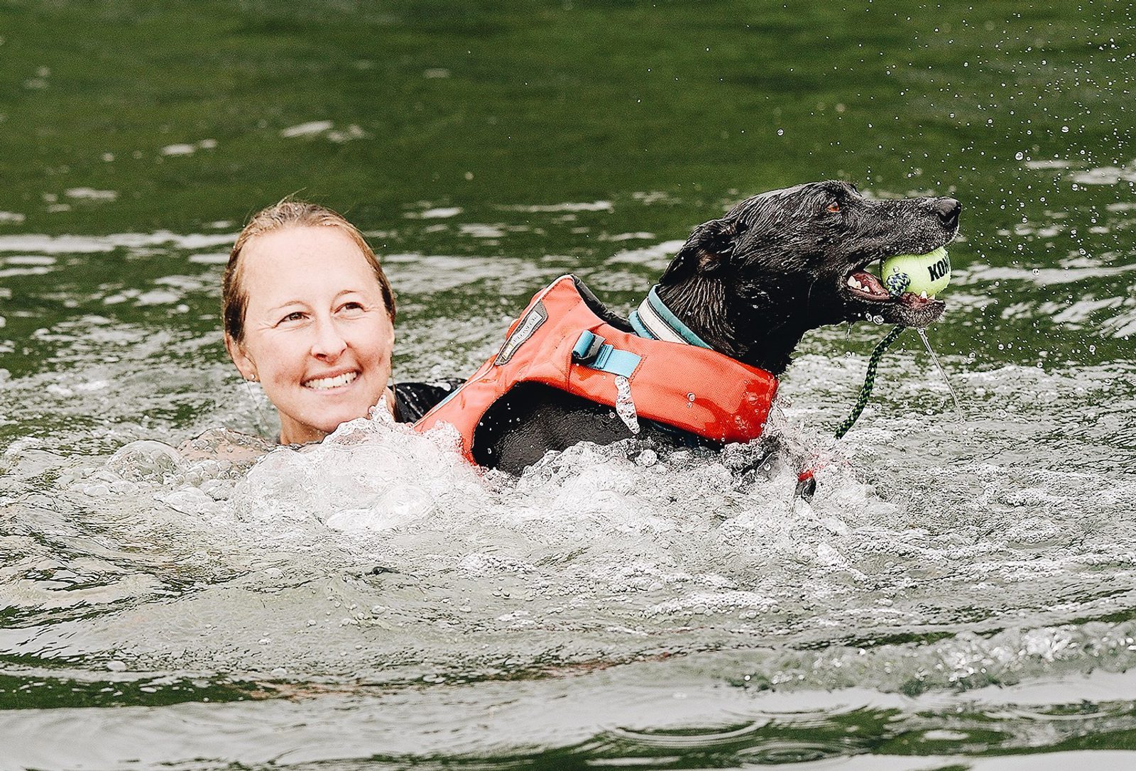 A woman smiles while swimming in a body of water with a black dog. The dog, wearing an orange life vest, holds a tennis ball in its mouth. The water around them is rippling, suggesting movement and playfulness.