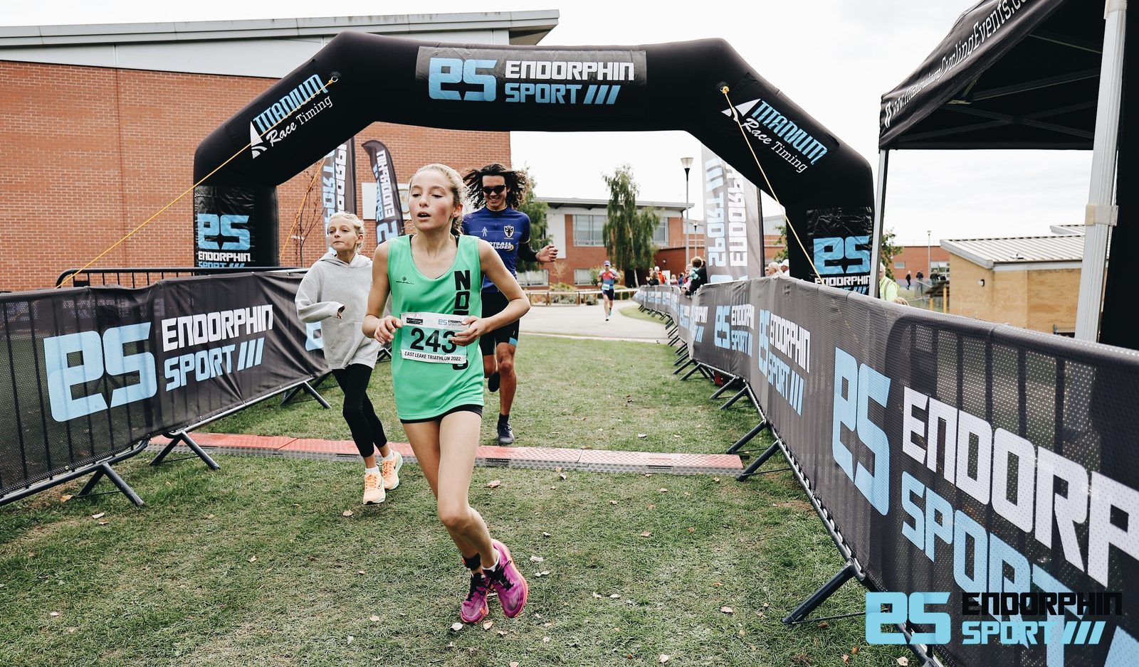 A young runner in a green uniform with the number 245 crosses the finish line at an outdoor race event. Others follow close behind her. The event is sponsored by Endorphin Sport as indicated by the banners and archway framing the finish line.