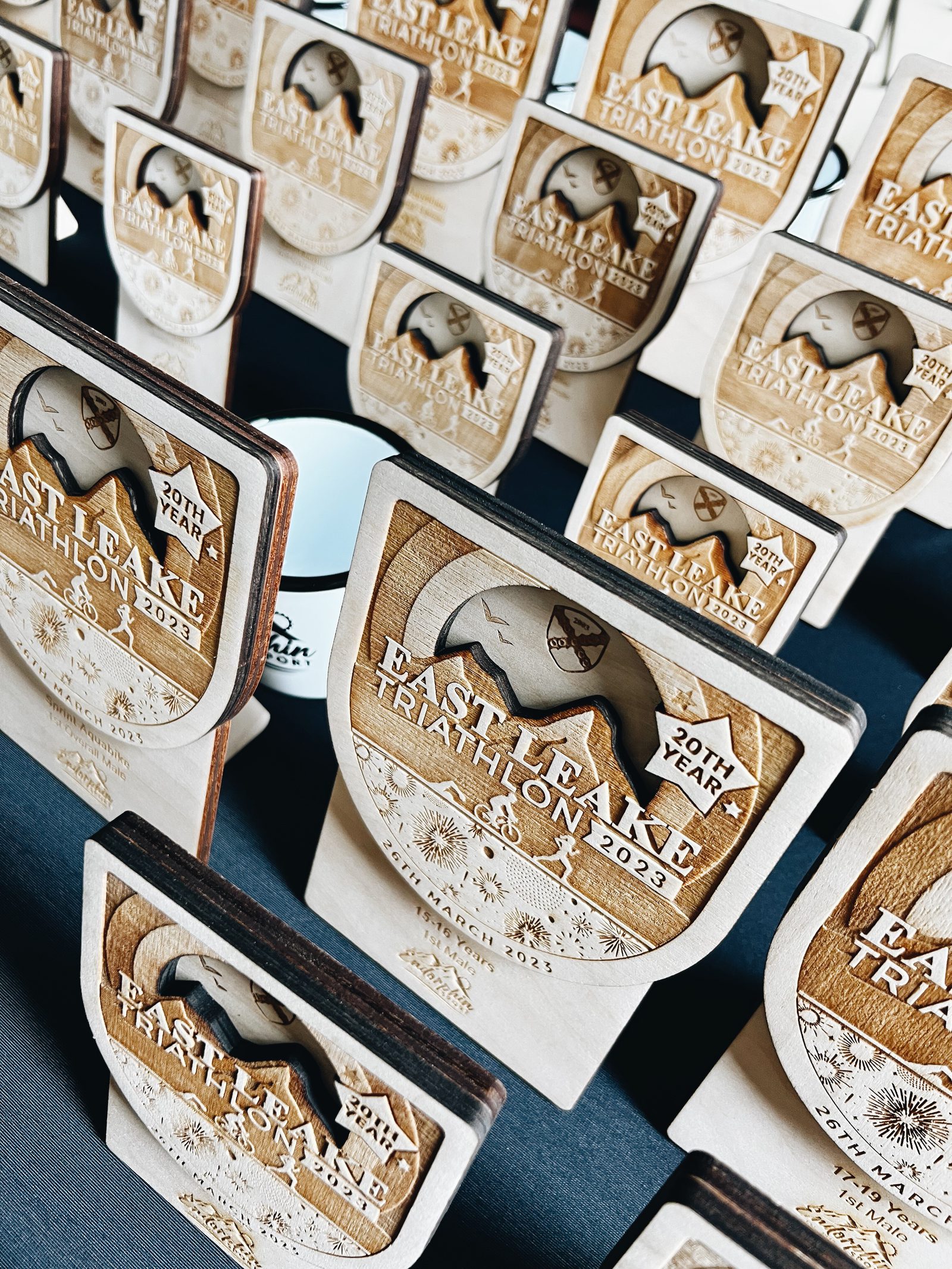 A close-up photo of several wooden awards featuring intricate designs. The awards, engraved with "Eastlake Triathlon" and the year "2023", commemorate the 20th year of the event. The awards display a sun and waves motif, arranged neatly on a dark surface.