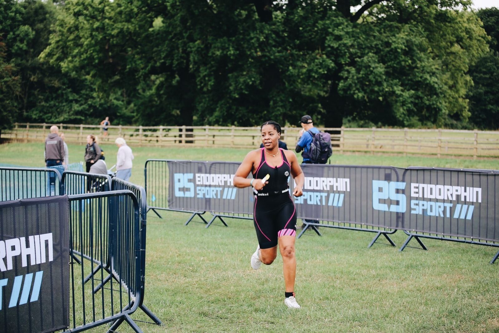 A female athlete in a black triathlon suit is running on a grassy path. She has a determined expression, and spectators are visible in the background. Barriers with the "ENDORPHIN SPORT" logo line the path, and trees provide a greenery backdrop.