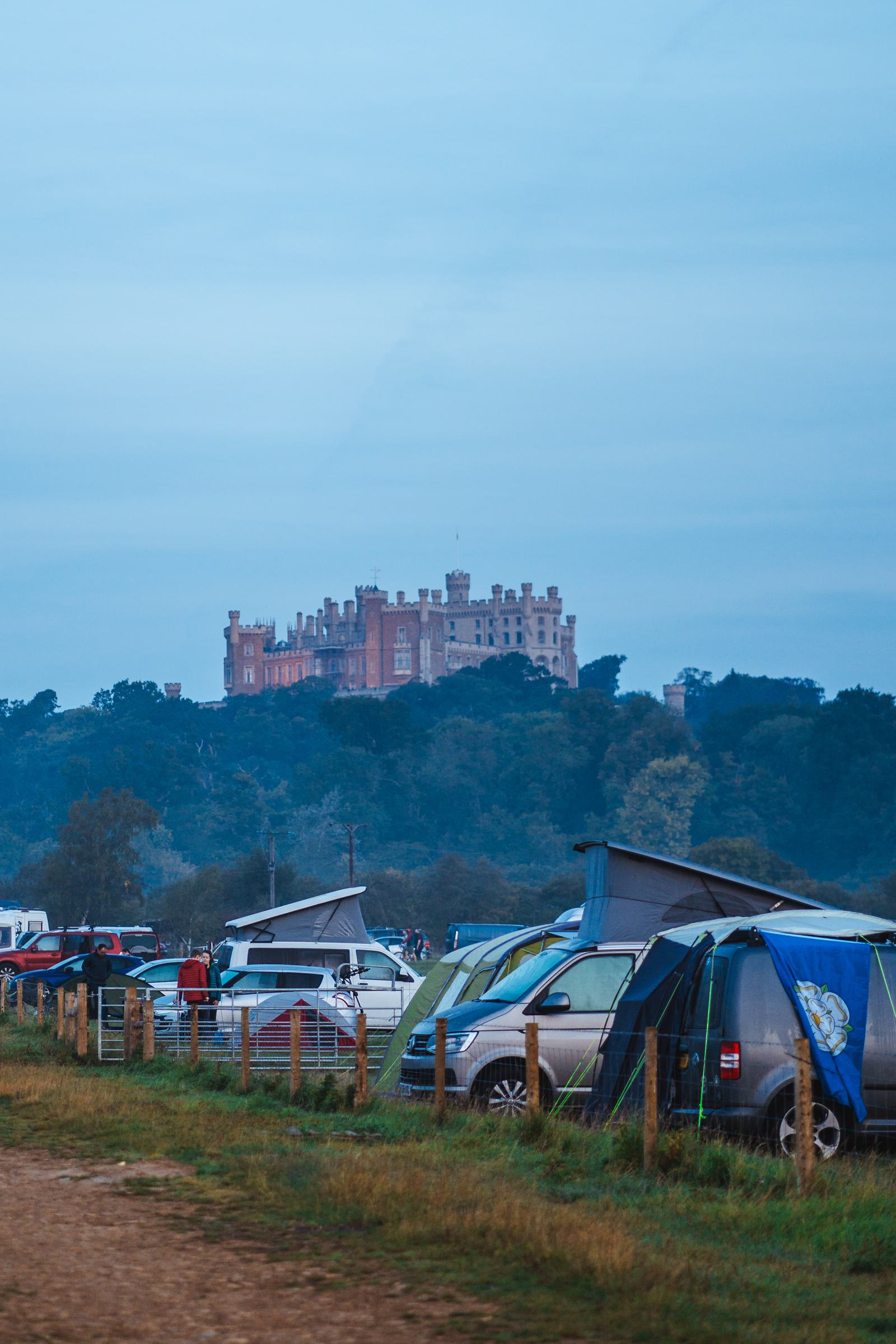 A large castle sits atop a hill surrounded by trees under a cloudy sky. In the foreground, there are several tents and camper vans in a grassy area, with a dirt path running along the side.
