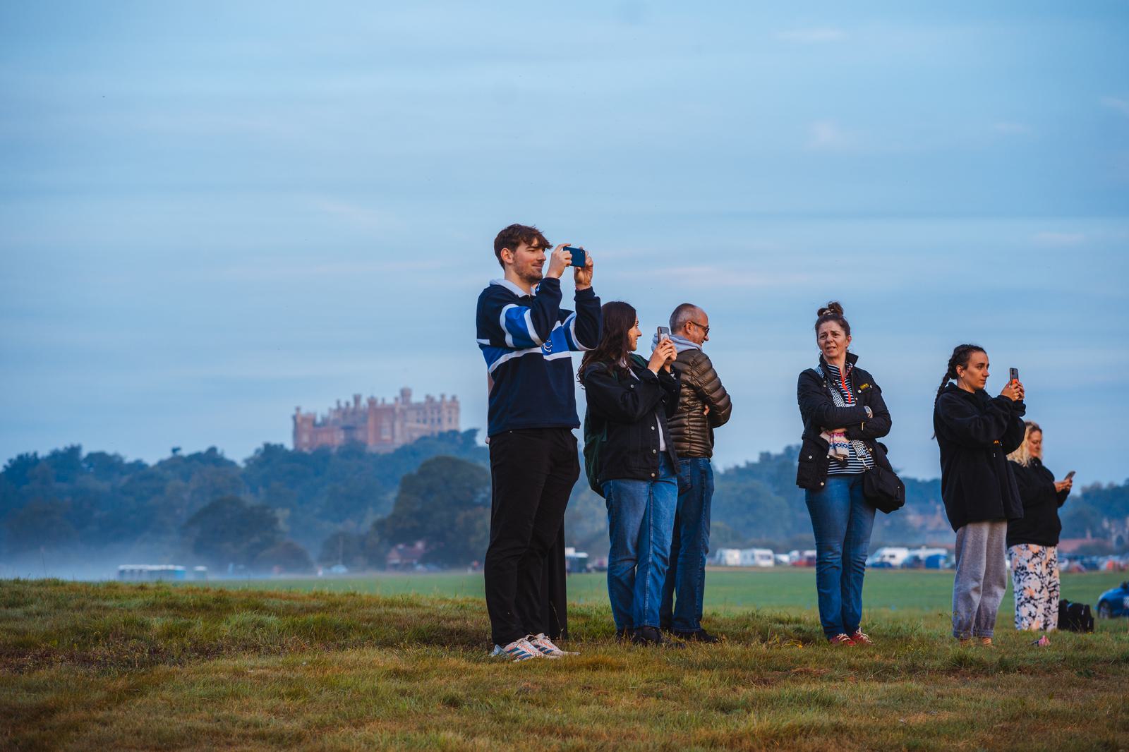 A group of people stand on a grassy field, some holding cameras and phones, capturing a distant castle in the early morning light. The sky is partially cloudy, and a light mist covers the background.