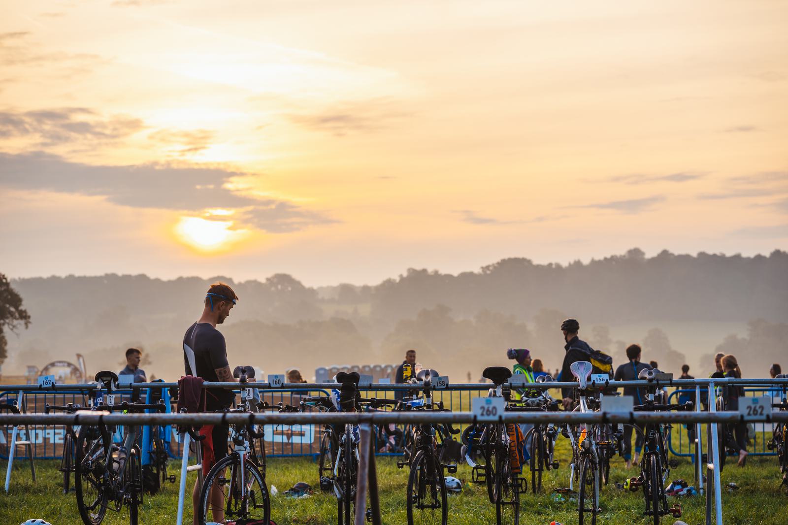 Cyclists preparing their bikes for a race at sunrise, with a scenic background of rolling hills and a vibrant sky. Racks labeled with numbers are visible, and participants are wearing athletic gear.