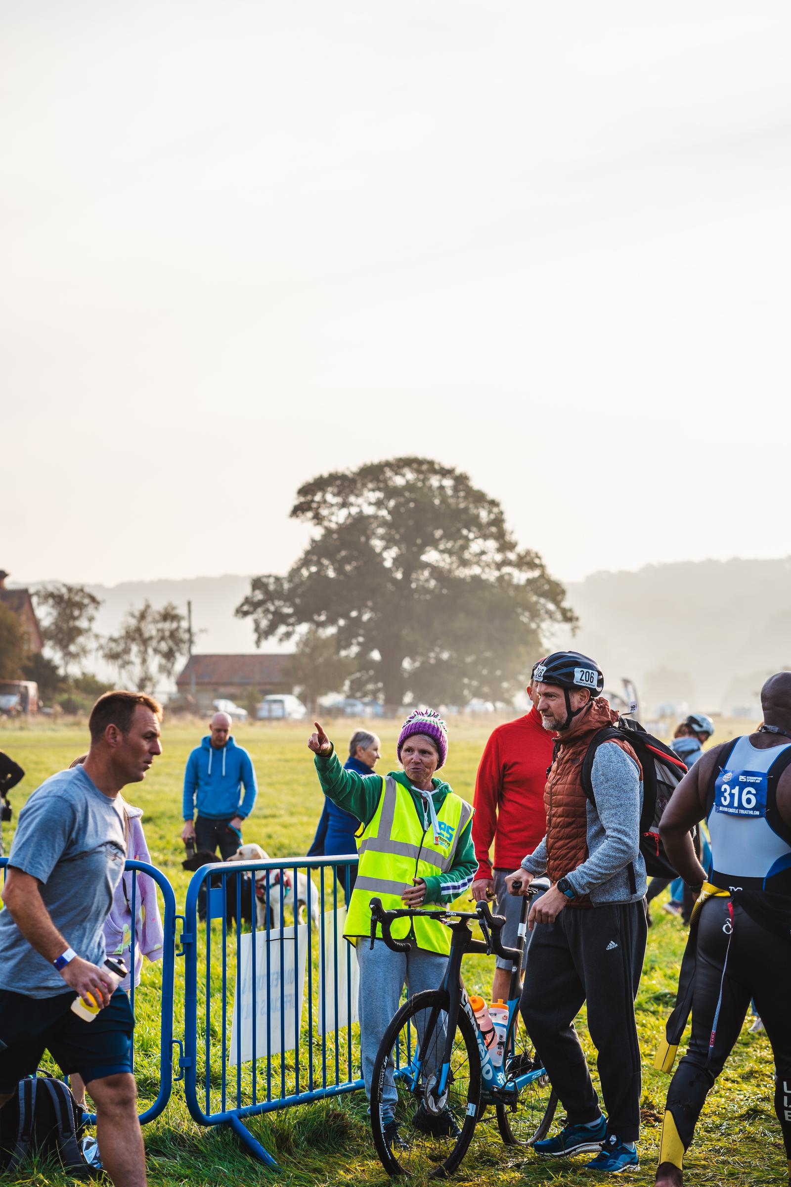 People gather outdoors at a sporting event. A person in a high-visibility vest points while others, some with bicycles, listen. The setting is a grassy field with trees in the background.