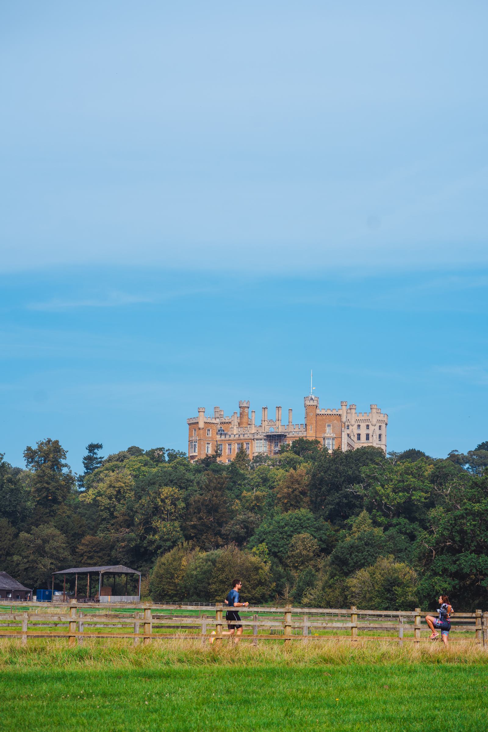 A scenic view of a large castle with numerous turrets, situated on a hill surrounded by lush green trees. Two people are walking on a path in the foreground, with a wooden fence and a grassy field separating them from the castle.