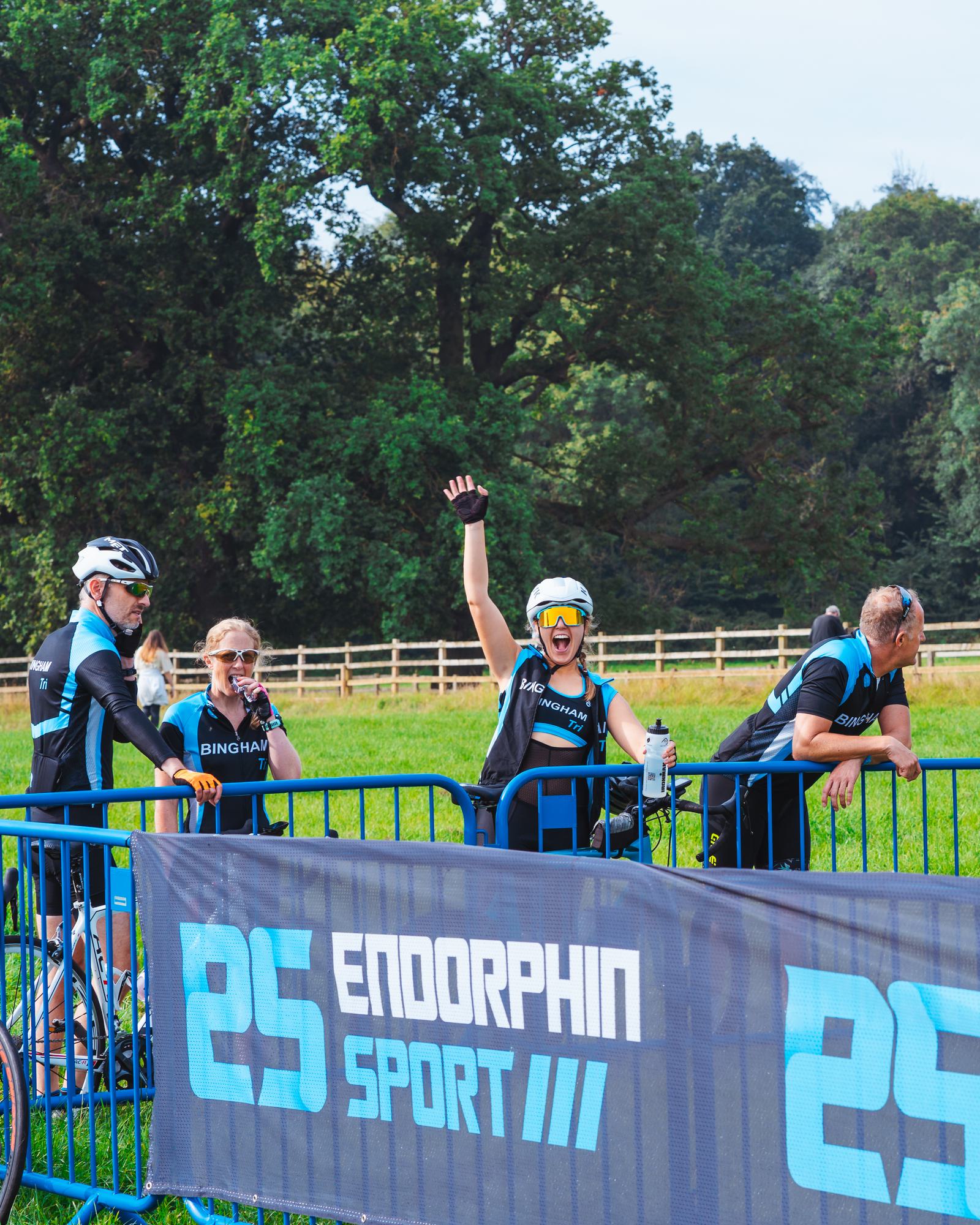Cyclists in matching black and blue jerseys stand near a fence with "Endorphin Sport" printed on it. One cyclist waves energetically at the camera. Green trees and a fence line form the background.