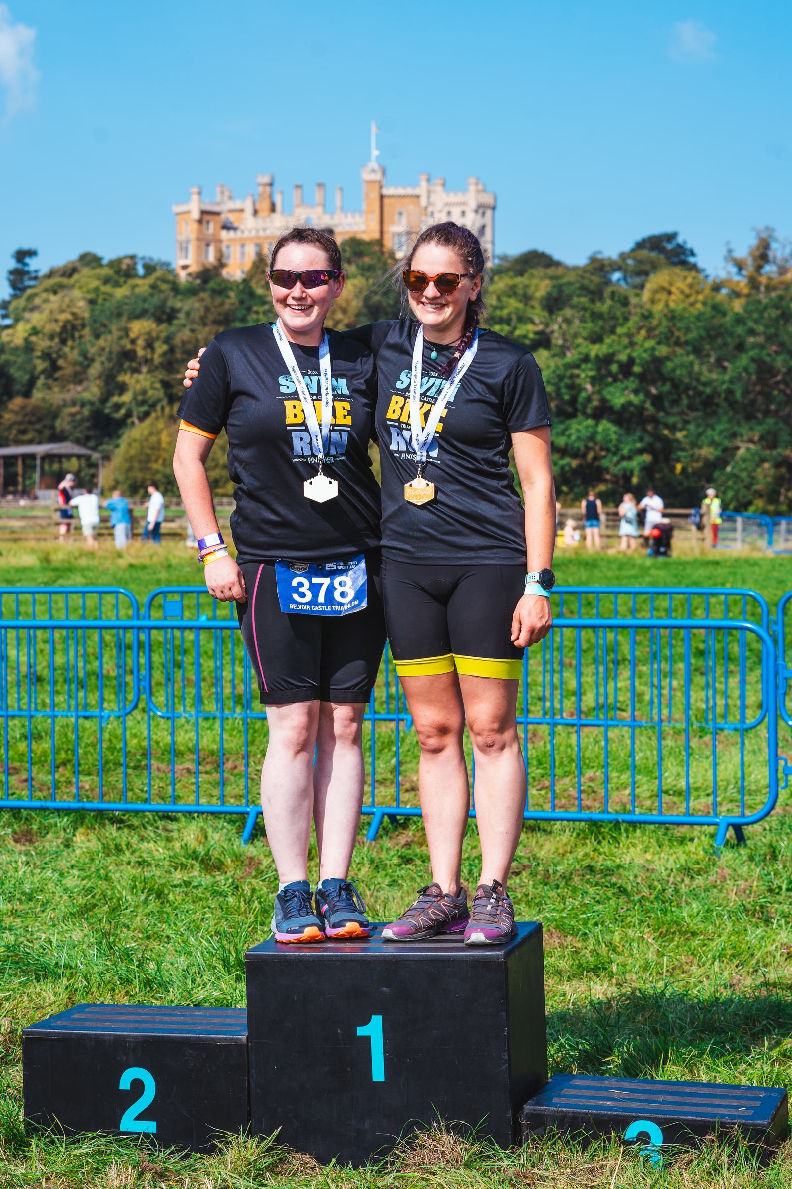 Two women standing on a podium after a bike ride event, wearing medals and black shirts with "Bike Ride" text. They're smiling, with a large building and trees in the background. One stands on the first-place spot, the other on the second.