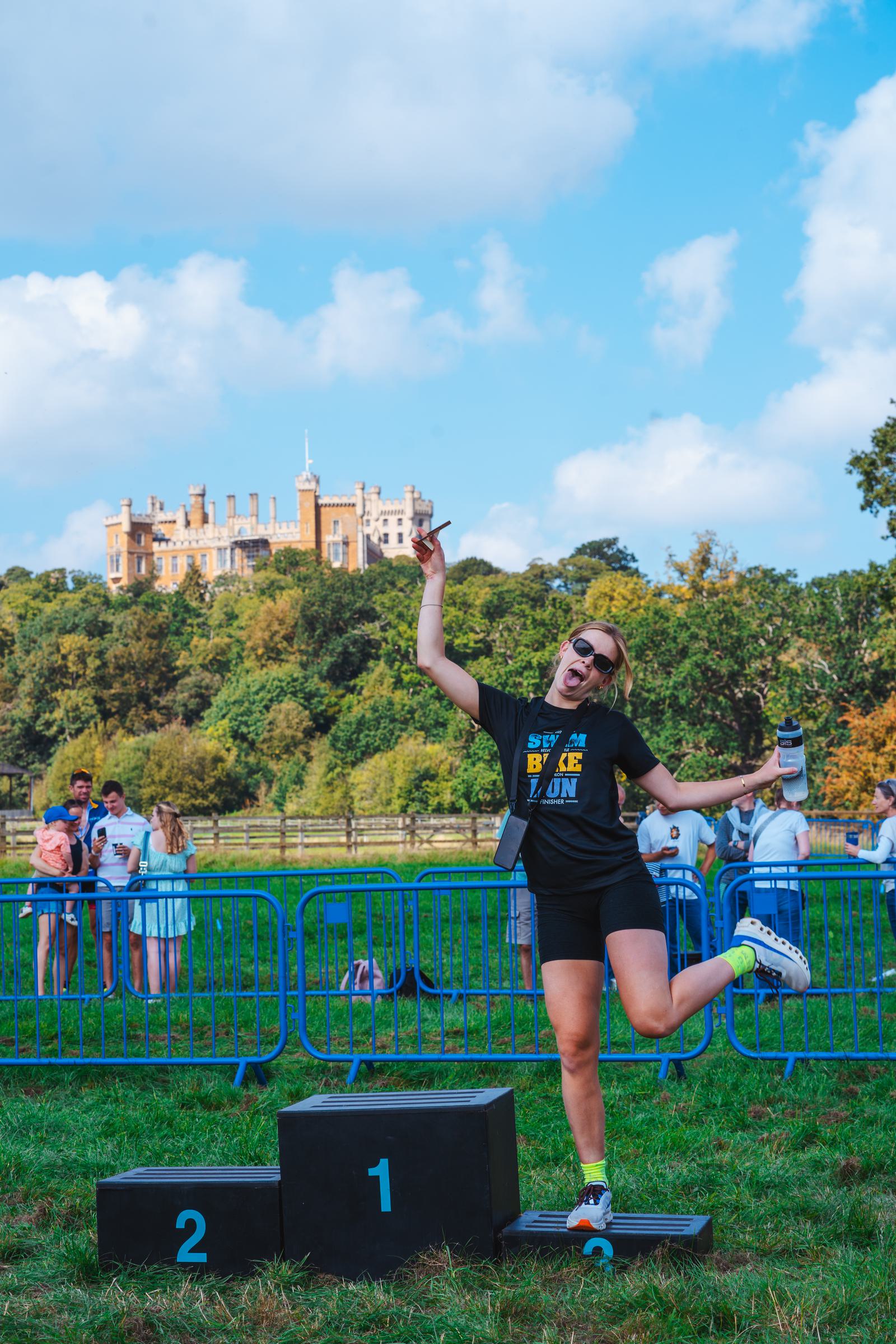 A person stands joyfully on a winner's podium, posing with a playful gesture. They are outdoors, with a historic building in the background. The person is wearing sunglasses and holds a bottle, surrounded by greenery and a blue sky.