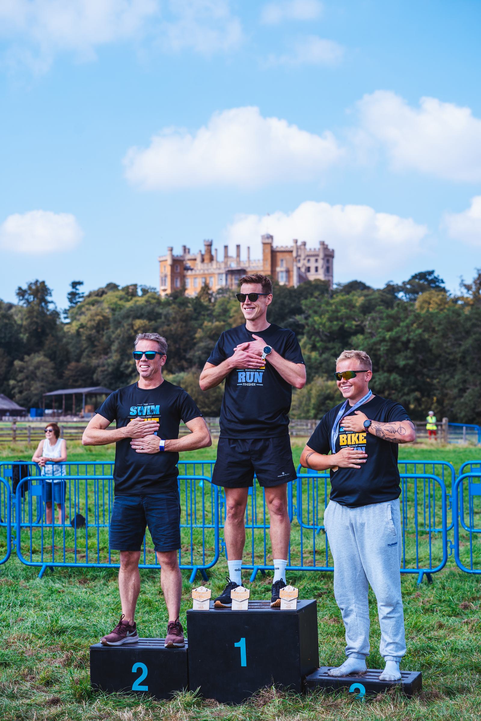 Three men stand on a podium in an outdoor setting, each wearing themed shirts for swim, run, and bike. The podium is numbered 1, 2, and 3. A castle and trees are visible in the background under a blue sky.