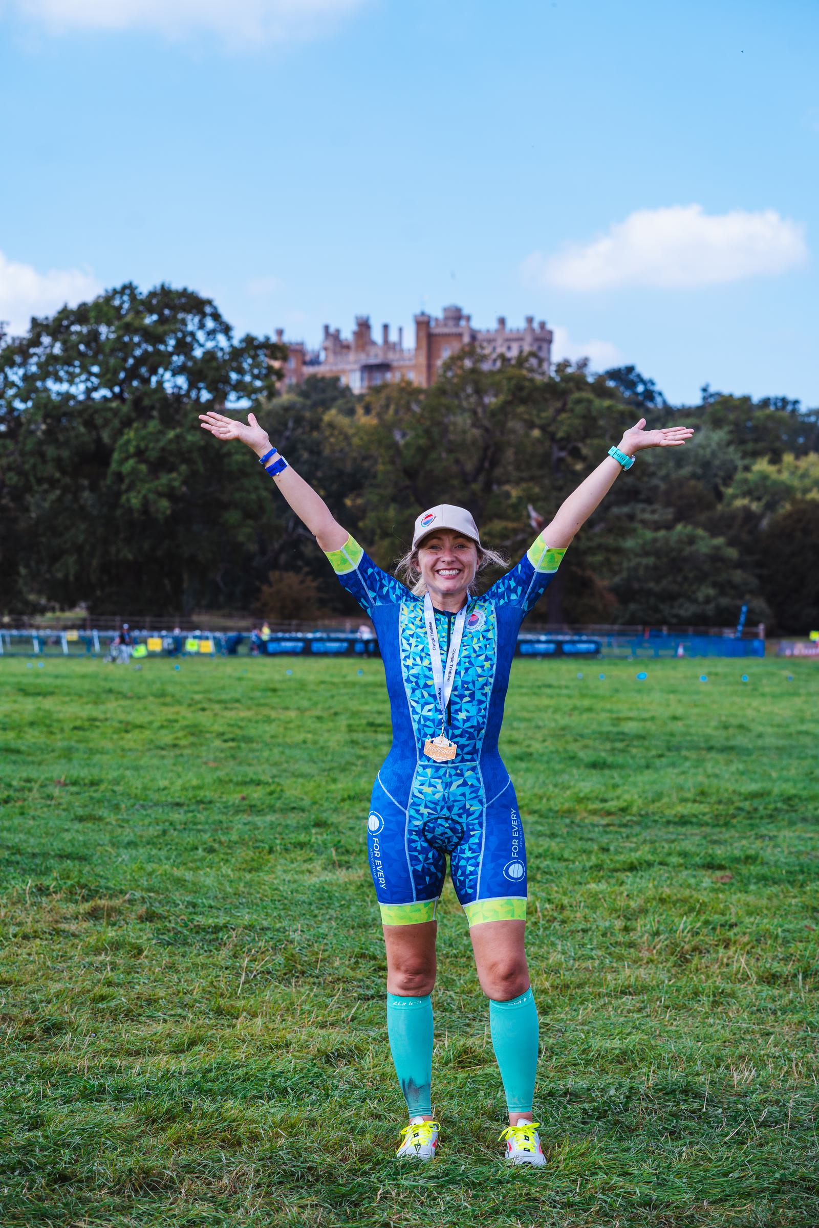 A person stands proudly on a grassy field with arms raised, wearing a medal and a colorful athletic outfit. A cap shades their face, and a castle is visible in the background under a blue sky.