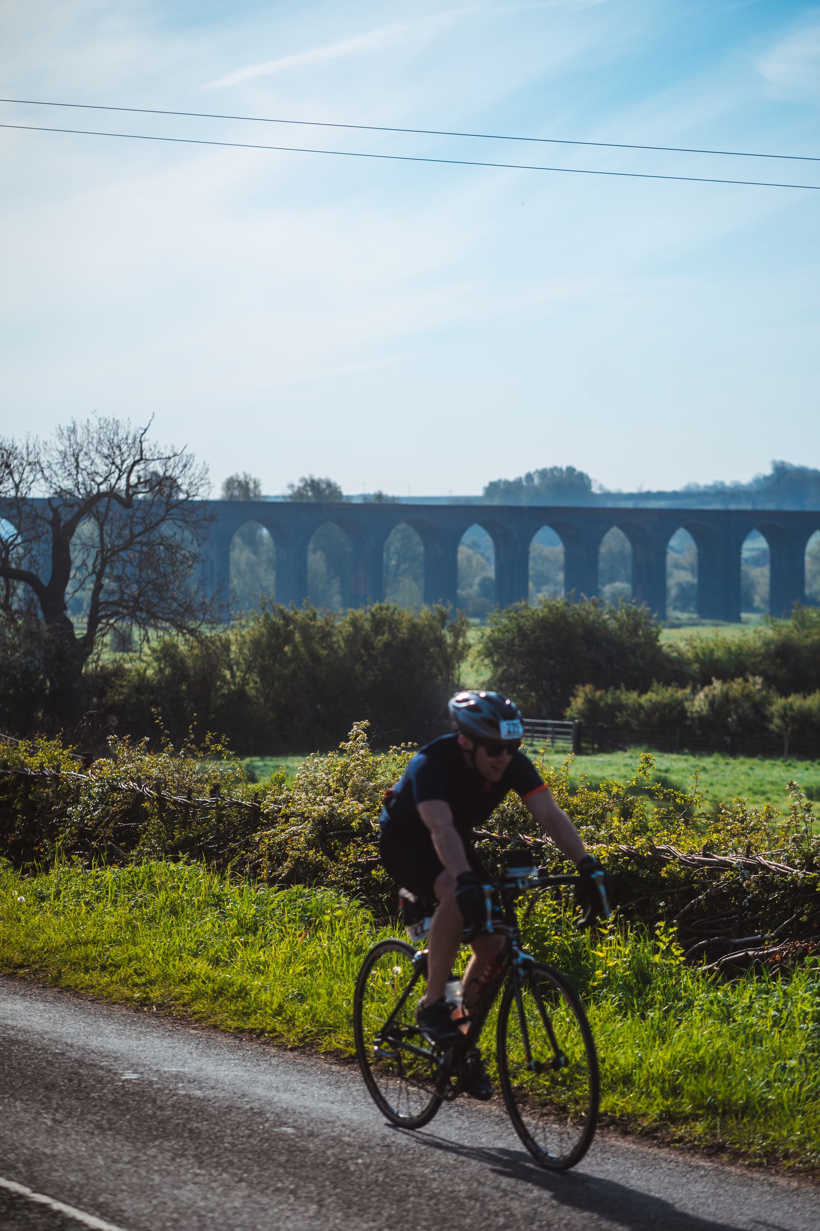 A cyclist wearing a helmet rides along a rural road, with a tall stone viaduct and lush green countryside in the background under a clear blue sky.