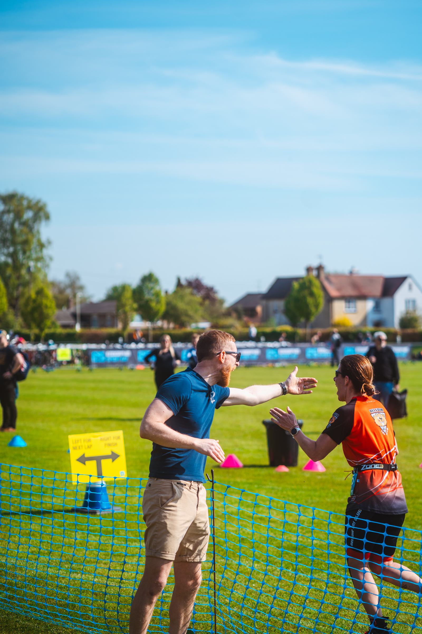 A man in a blue shirt gestures animatedly towards a woman in an orange shirt at an outdoor event. They stand near a blue mesh fence on a grassy field under a clear blue sky. Houses and people are visible in the background.