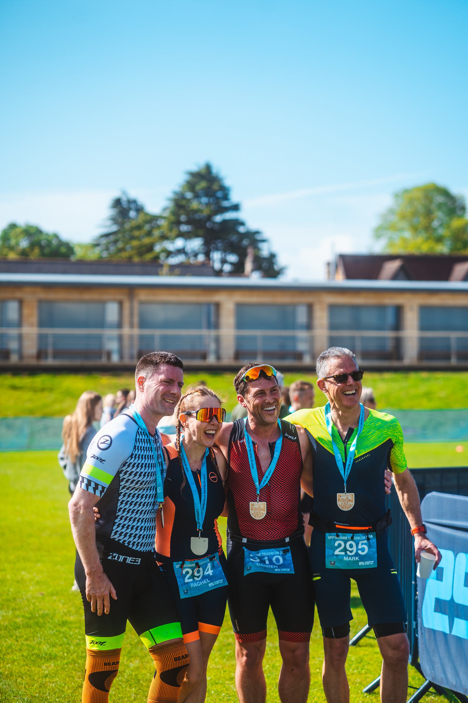 Four athletes in sporting attire stand together on a grassy field, smiling and displaying medals around their necks. They are celebrating their achievement with arms around each other. In the background, a building and trees are visible.