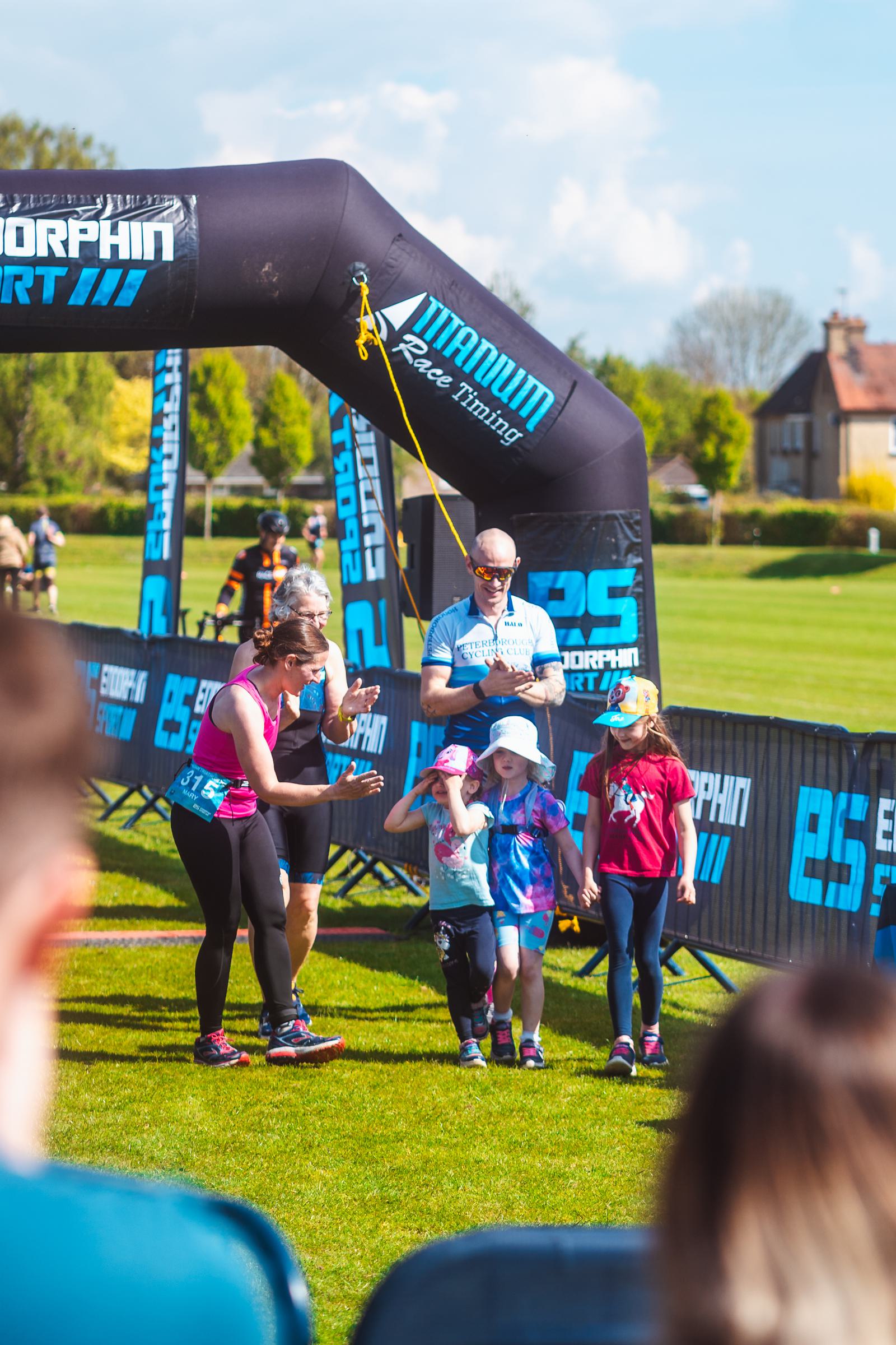 A group of people, including adults and children, walk under a race finish arch outdoors. The scene is sunny, with blue banners and lush greenery in the background. The children are smiling and wearing colorful clothes.