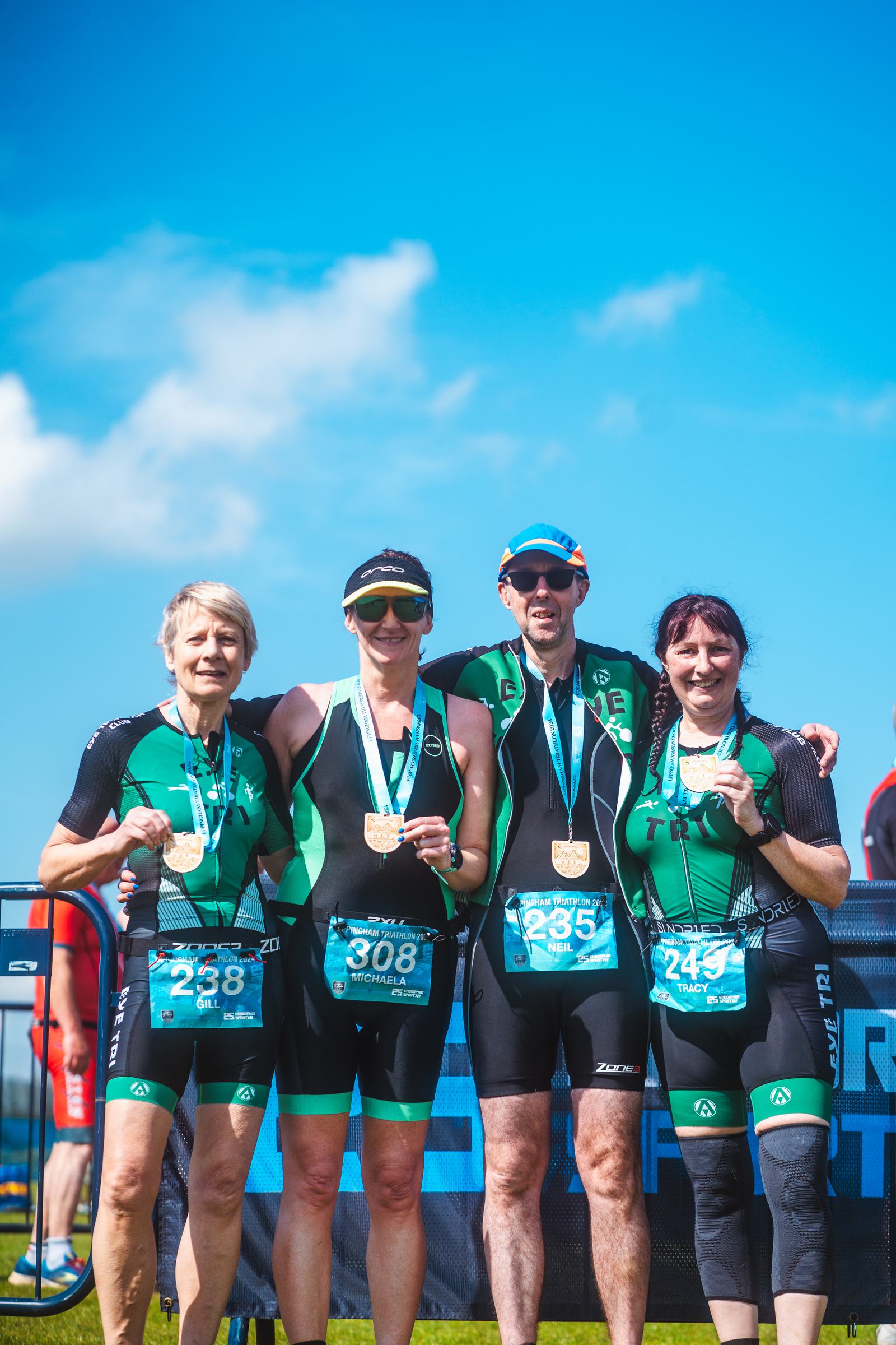 Four athletes wearing medals pose together after a race. They are dressed in matching athletic gear and stand in front of a banner, smiling under a clear blue sky.