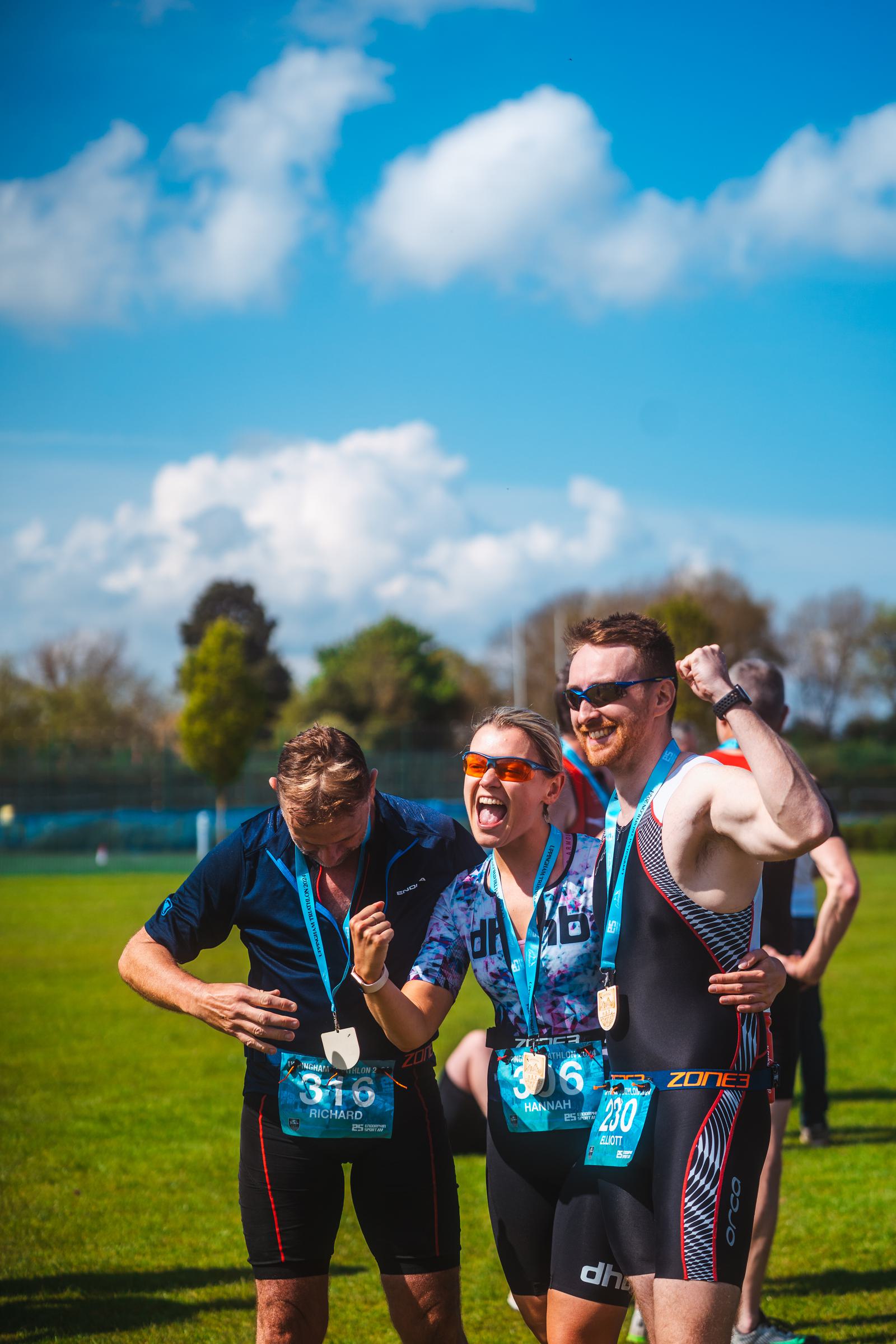 Three triathletes celebrate after finishing a race. They are wearing medals and athletic gear, smiling and posing triumphantly on a grassy field under a clear blue sky.
