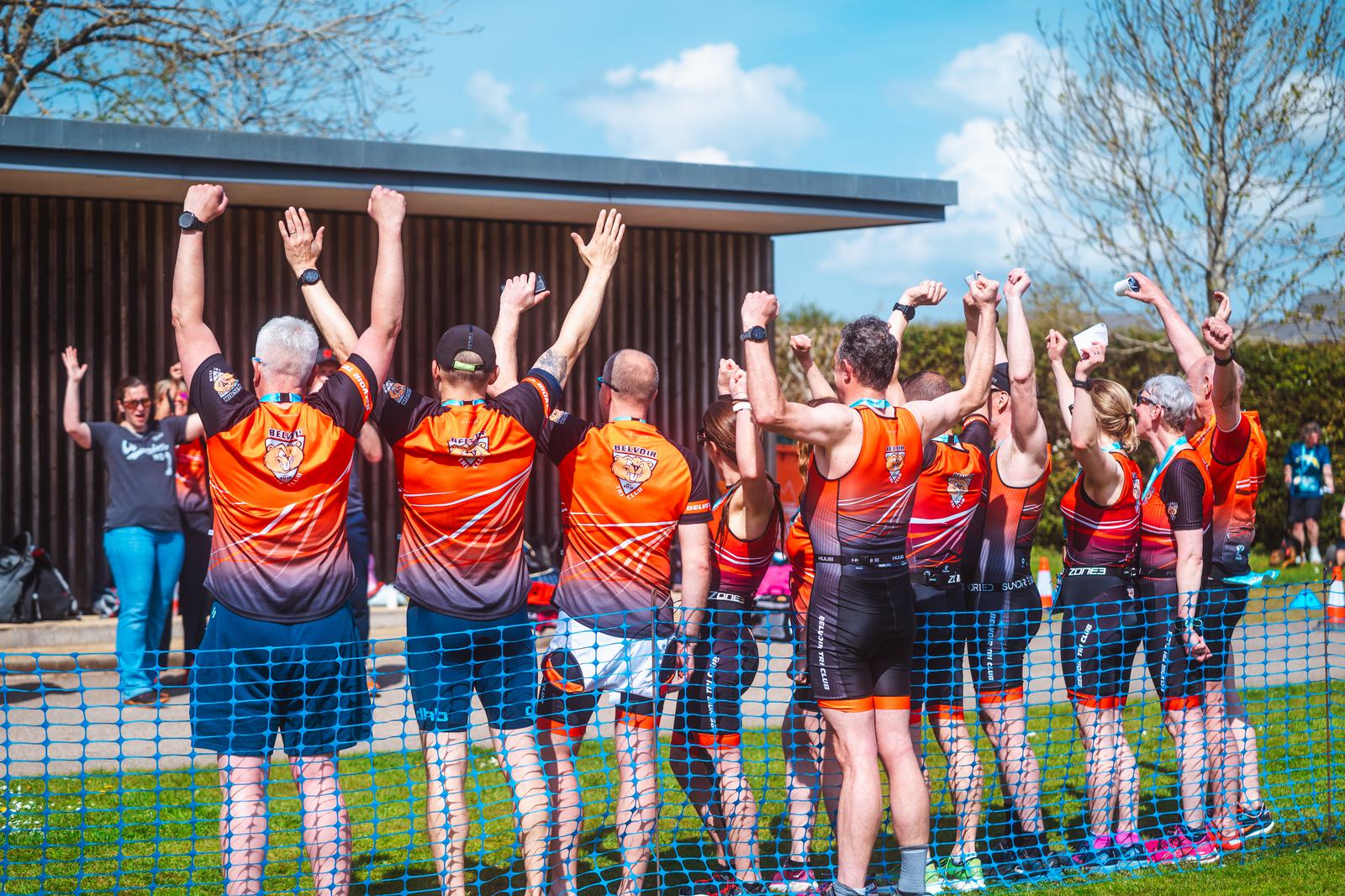 A group of athletes wearing matching orange and black sports attire stand with their backs to the camera, arms raised in celebration. They are on a grassy field near a building, with a blue net in front of them and a person applauding nearby.