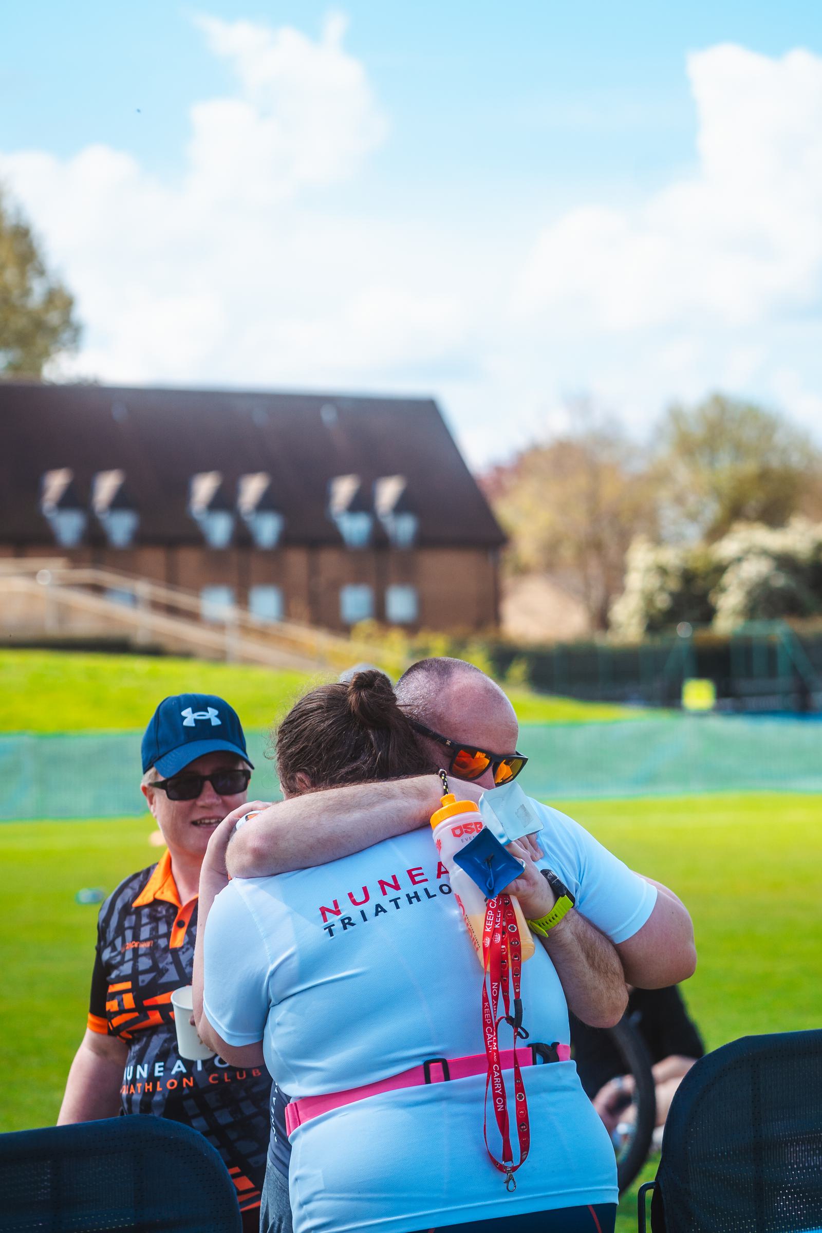 Two participants, wearing Nuneaton Triathlon shirts, hugging outdoors with medals around their necks. Another person in a cap stands nearby, smiling. Green field and trees in the background under a blue sky with clouds.
