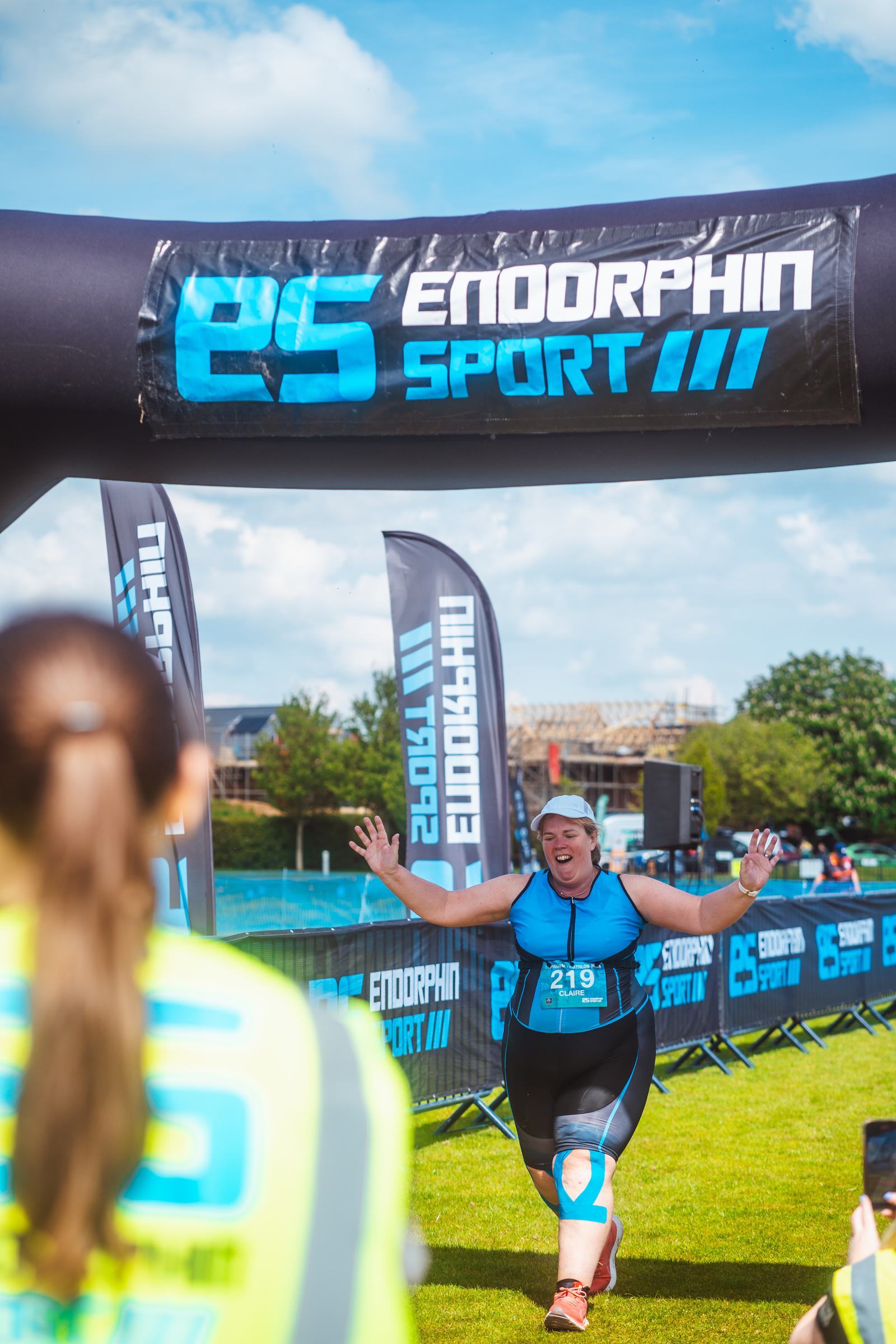 A person in athletic gear triumphantly crosses a race finish line with hands raised. The inflatable arch reads "Endorphin Sport." Spectators and banners are in the background on a sunny day.