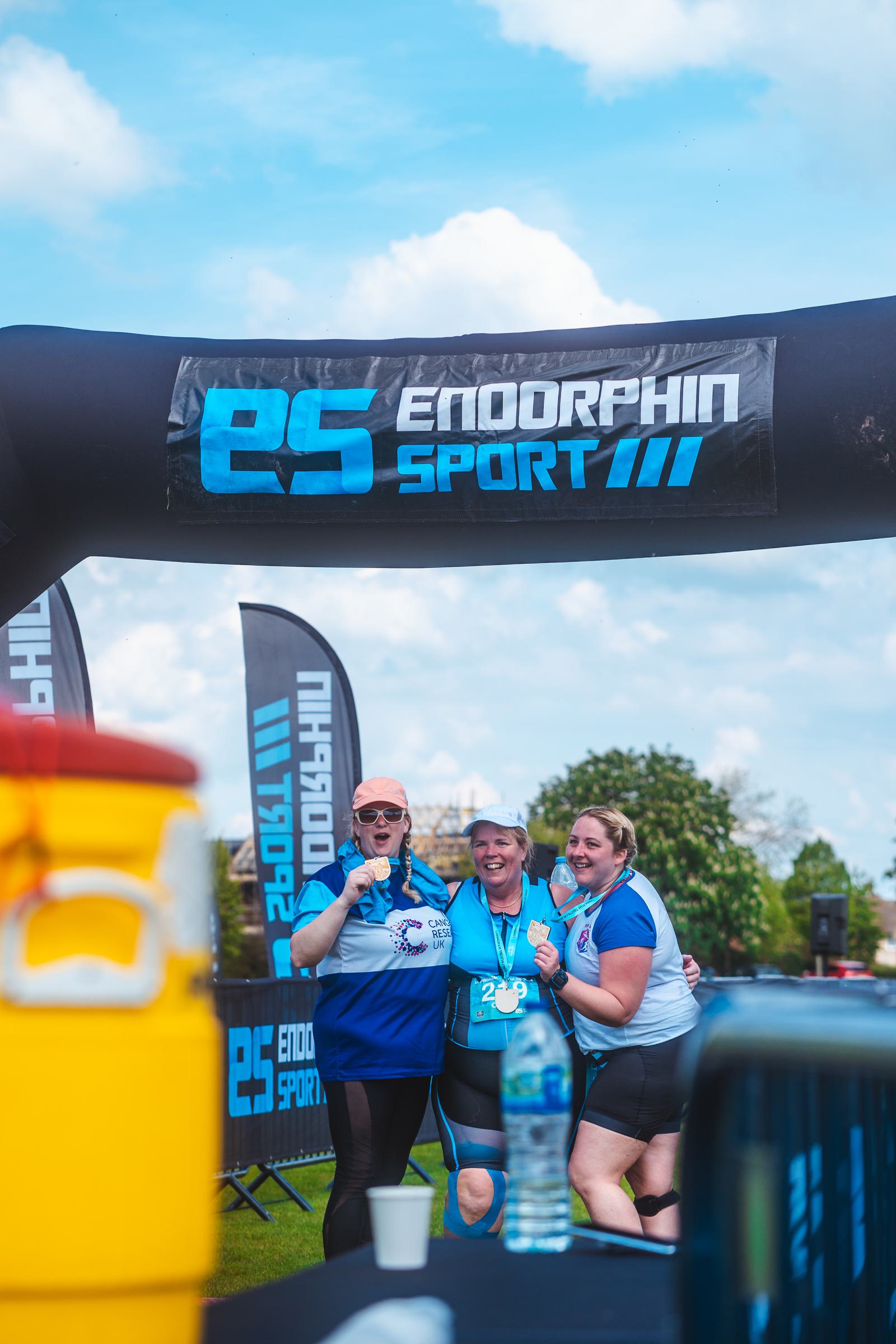 Three people in athletic gear celebrate under an inflatable arch that reads "Endorphin Sport." They hold finisher medals and pose for a photo. The sky is blue, and the setting appears to be an outdoor event.