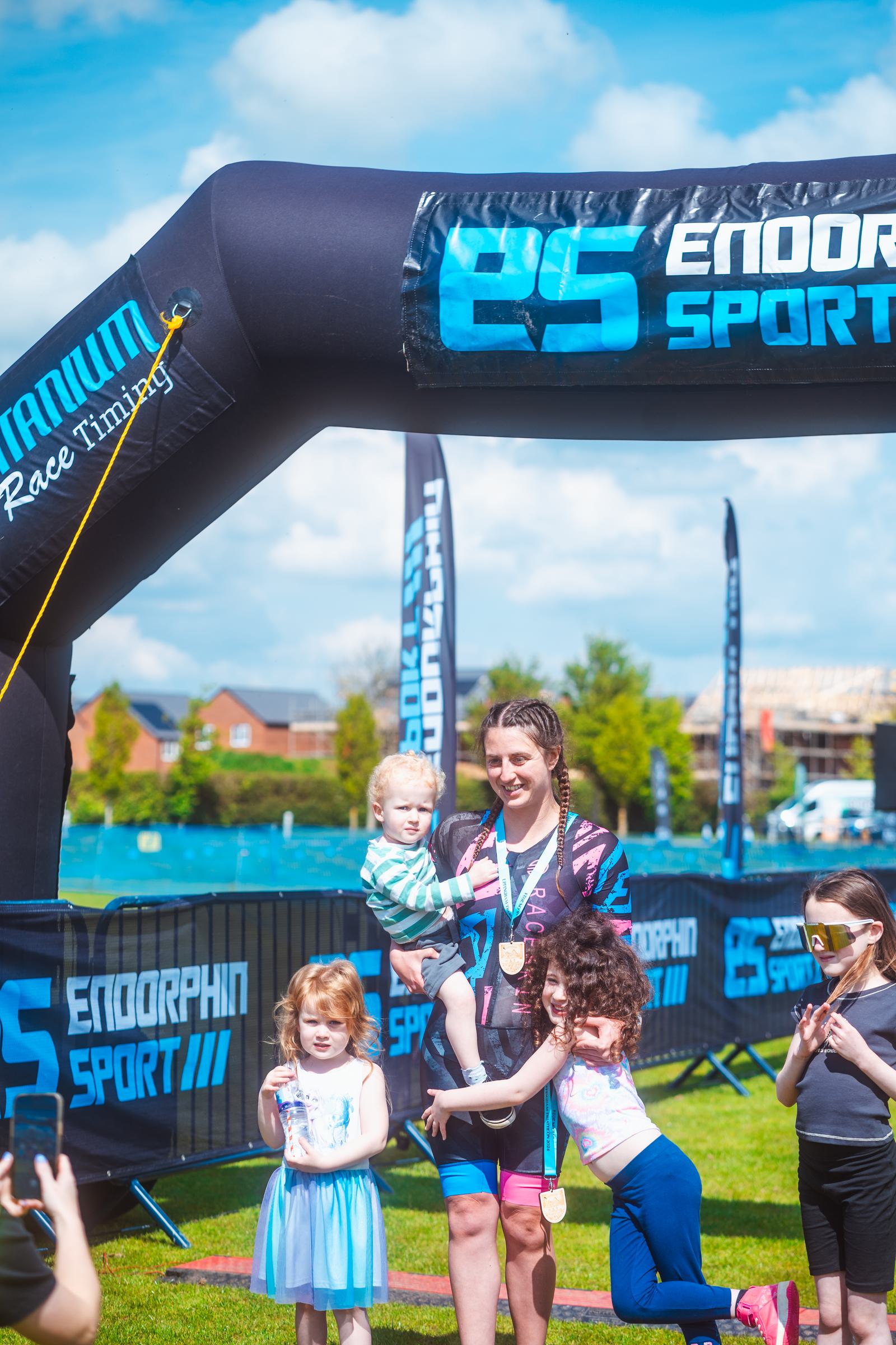 A woman stands under a "Endorphin Sport" finish line arch, holding a child. Two young girls stand beside her on the grass, with medals around their necks. Another person is to the side, wearing a mask. The sky is clear and sunny.