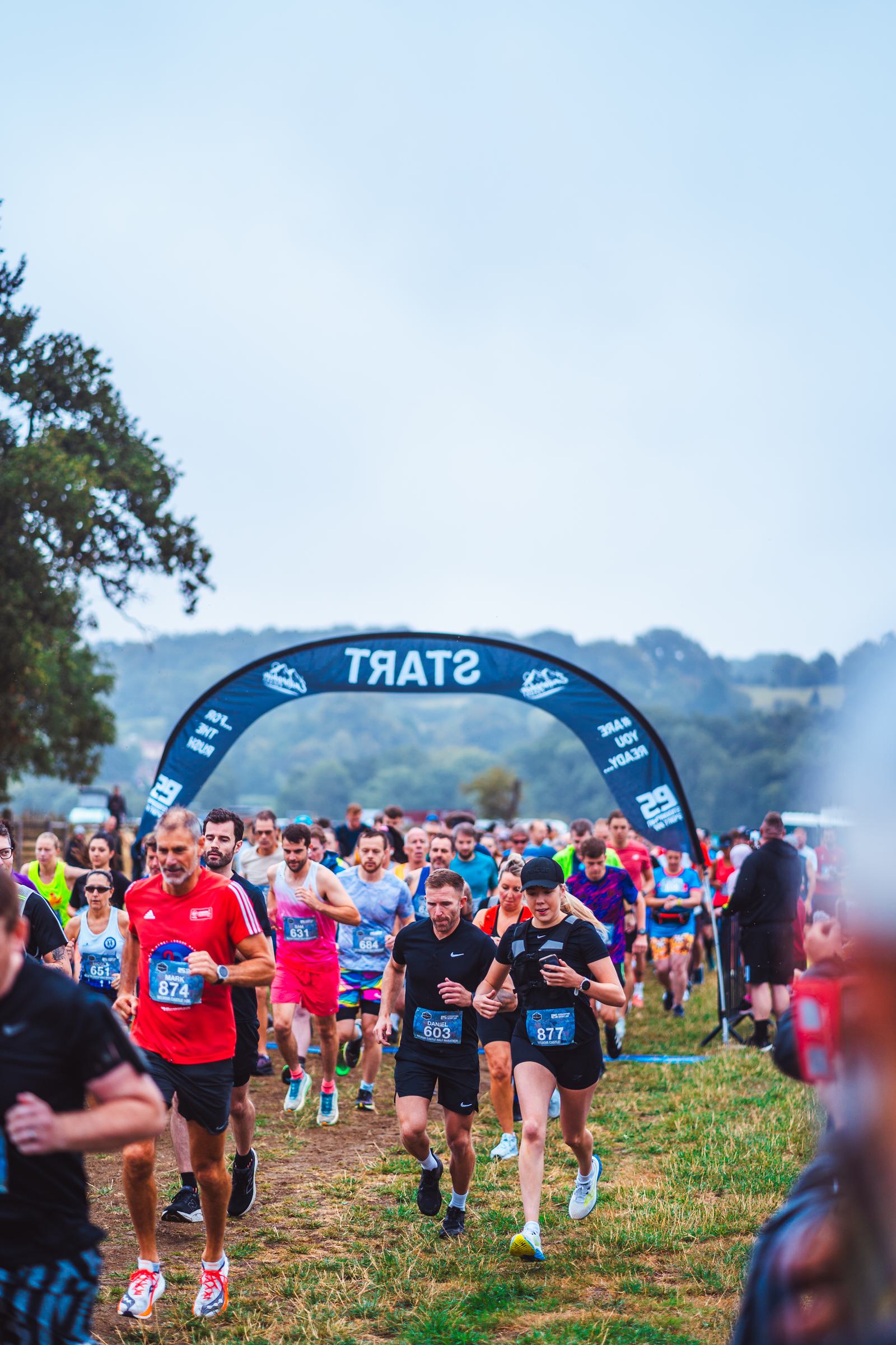 A group of runners participating in a race are passing under a blue arch labeled "START." The runners, wearing numbered bibs and athletic clothing, are on a grassy field with trees and hills in the background under a cloudy sky.