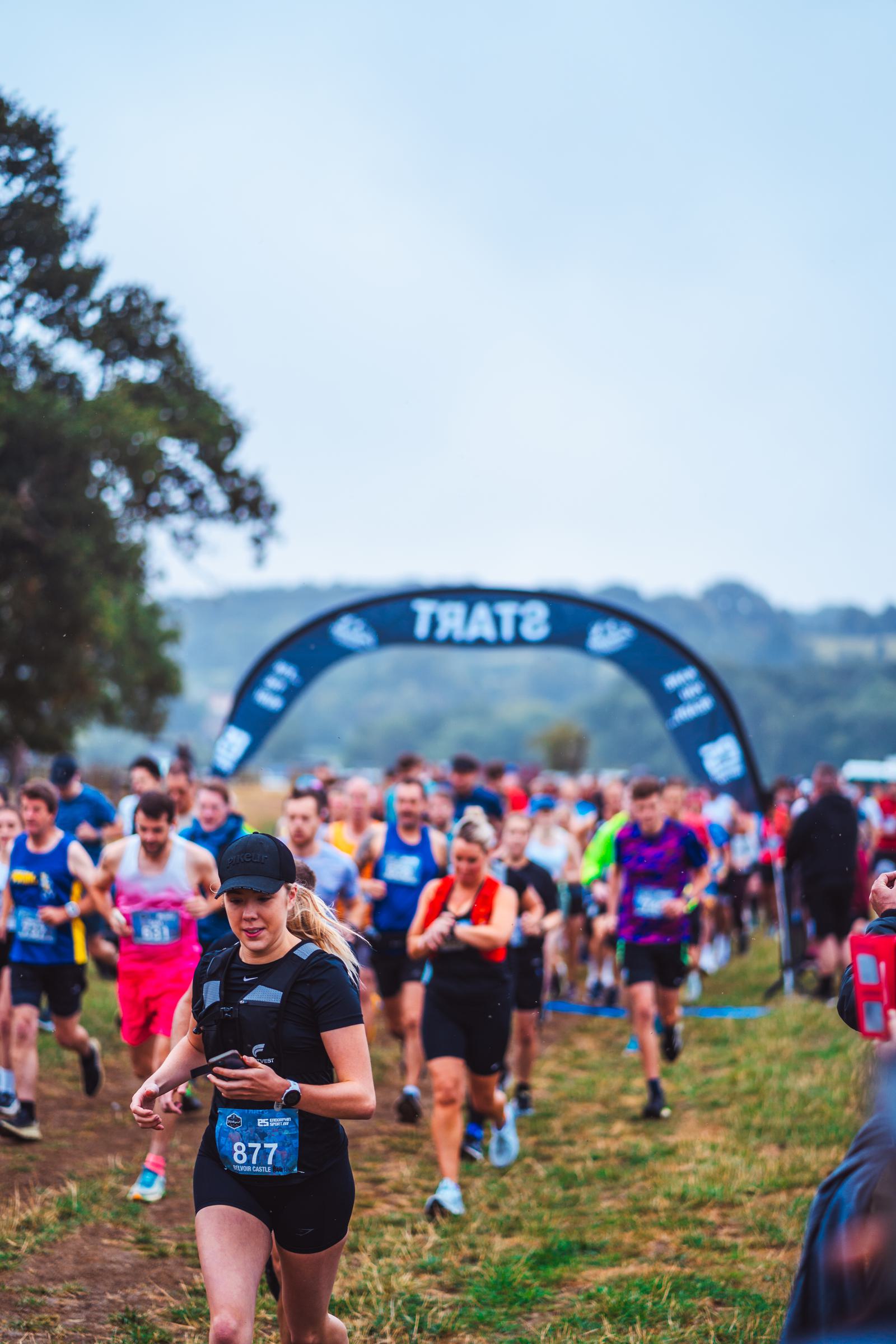 A large group of runners participating in a race. They are running on a grassy path with a blue "START" archway in the background. The sky is overcast, and trees are visible on the left side of the image.
