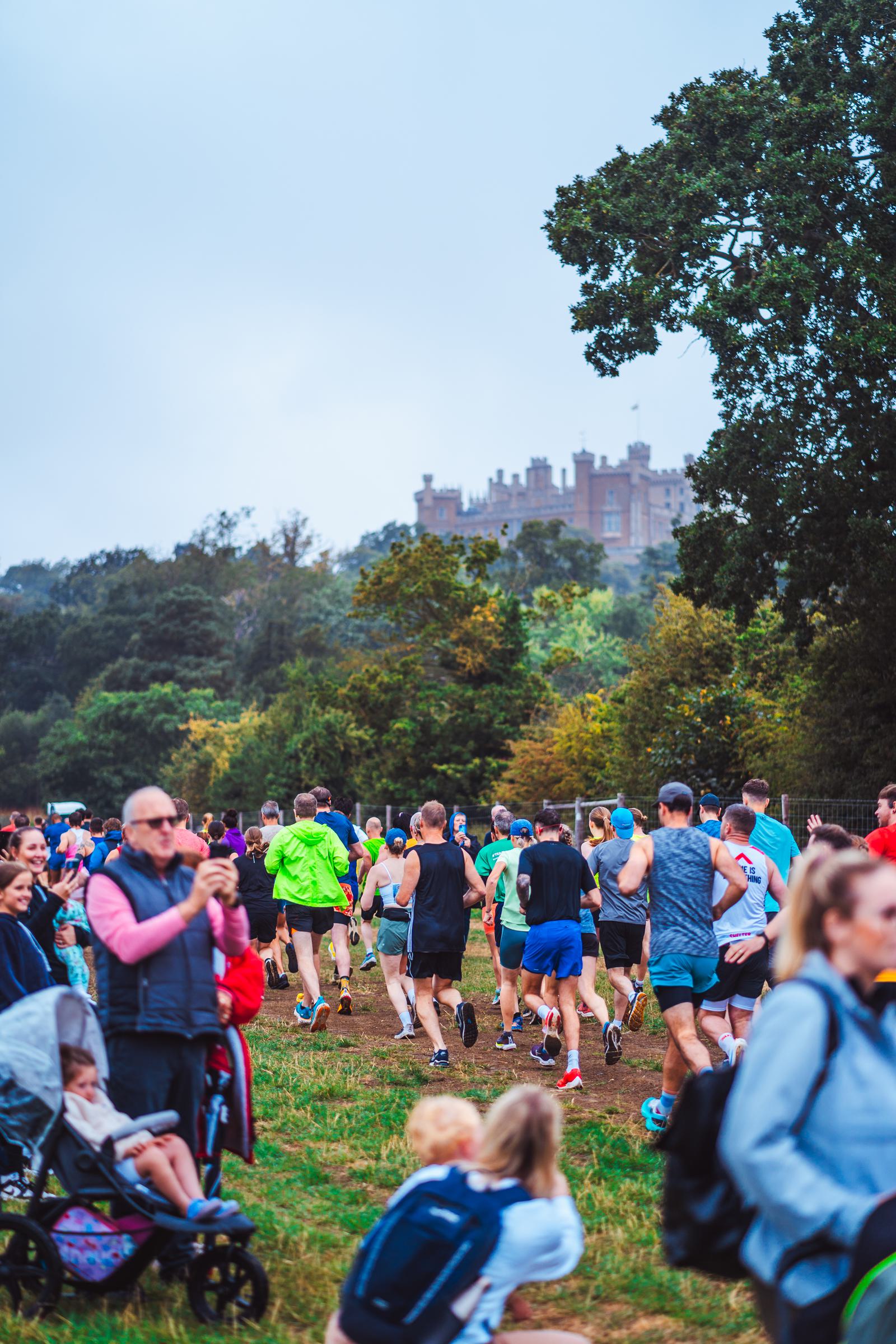 A group of people are jogging along a grassy path in a park. Spectators watch and take photos. A stroller and a child are visible in the foreground. Trees and a castle are in the background under a cloudy sky.
