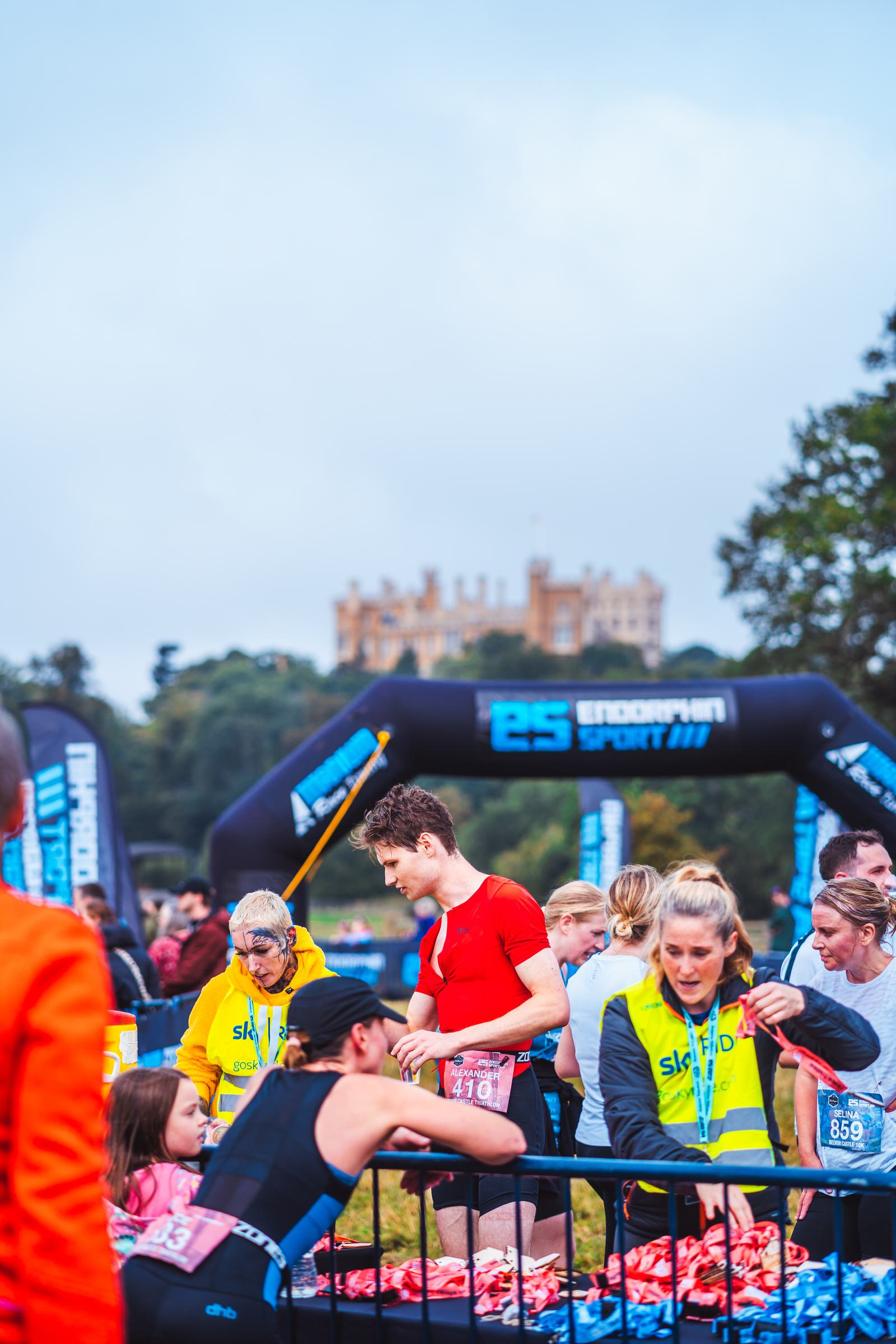 A group of people, wearing colorful athletic clothing, gather at a race or event. Some are checking their race numbers, while others are talking. A large inflatable arch is in the background, with a distant castle visible on a hill.