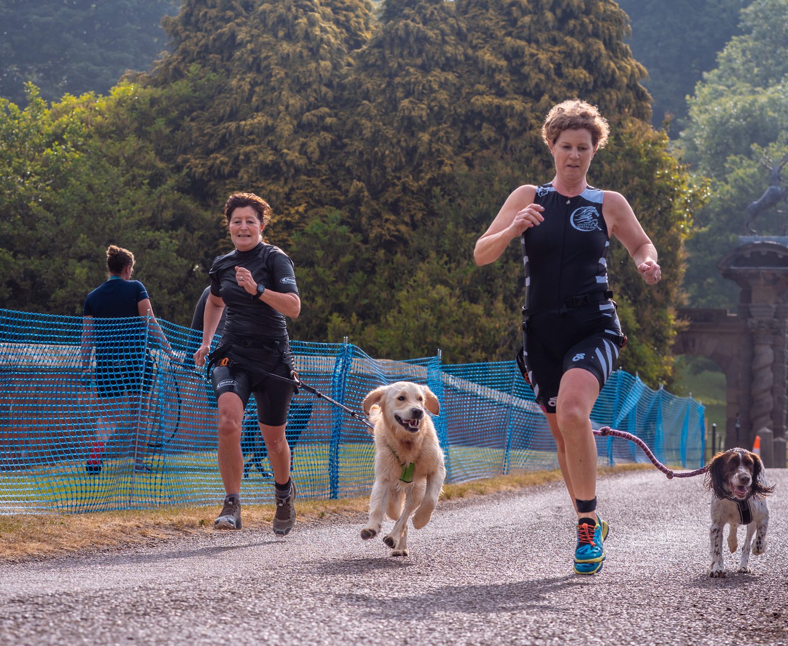 Two women running with dogs on a gravel path, surrounded by greenery. Both women wear athletic gear and each holds a leash; the one in front has a golden retriever, the other a smaller dog. Another runner is seen in the background, heading in the same direction.