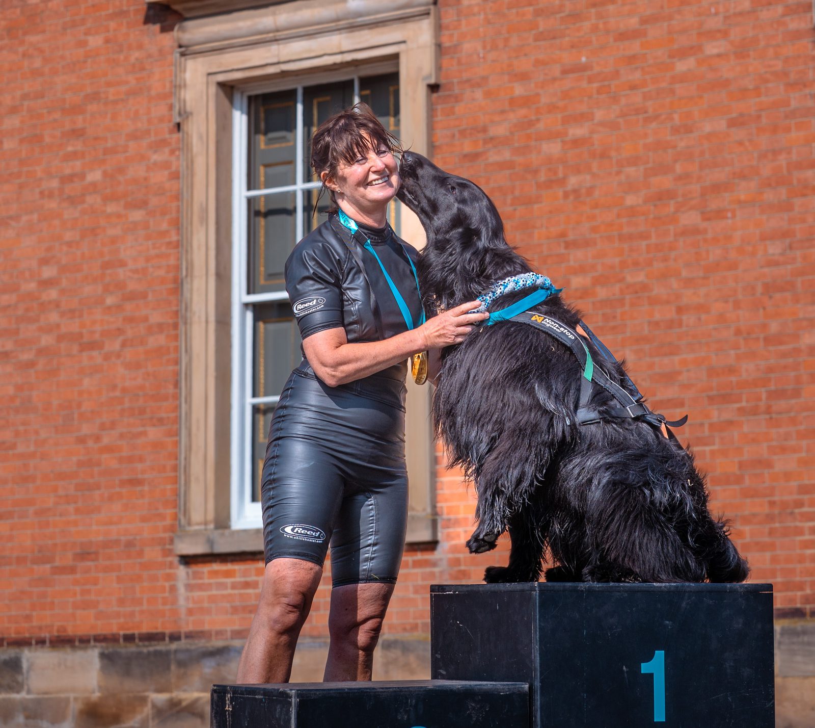 A woman in a wetsuit stands on a podium labeled "1" with a large black dog. The dog is licking the woman's face, and both wear medals around their necks. The background features a brick building with a window.