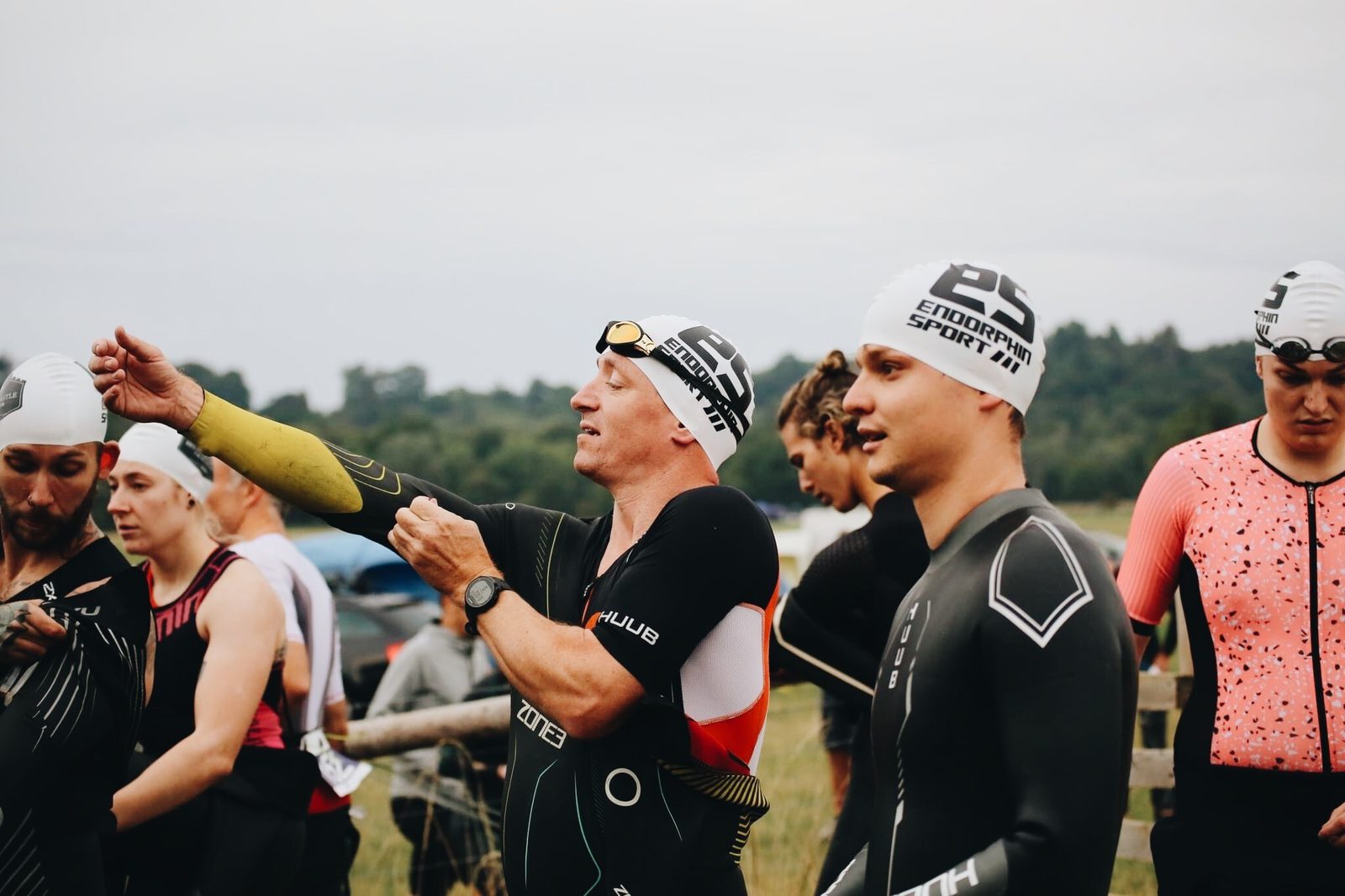 A group of triathletes prepares for a race, wearing wetsuits and swim caps, some branded with "Endorphin Sport." One man in the center adjusts his arm sleeve, while others focus on their own preparations. The background features a cloudy sky and trees.
