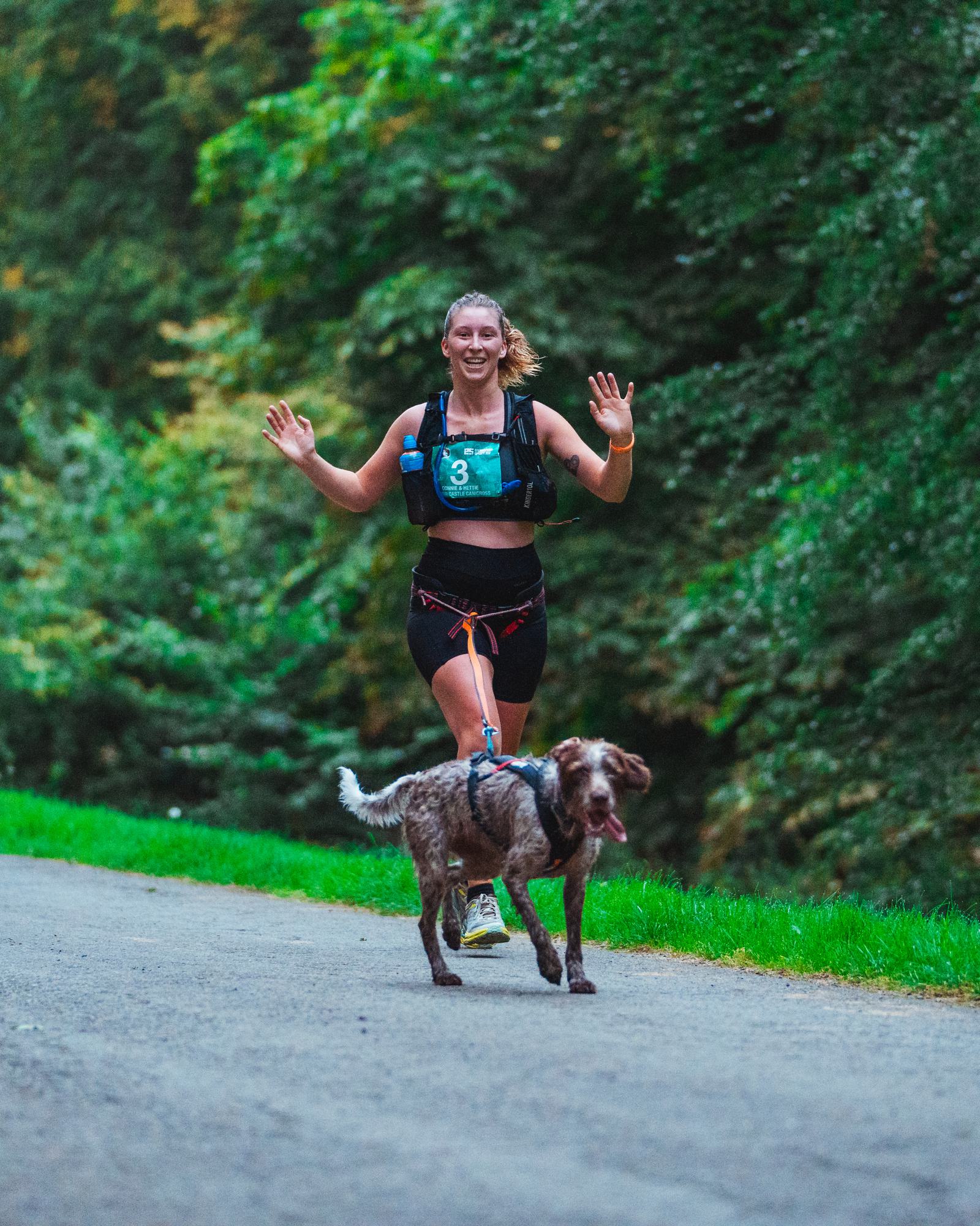 A woman is running on a paved path through a lush forest, smiling and holding her hands up in excitement. She is jogging with a dog attached to her waist harness, and both seem energetic and happy.