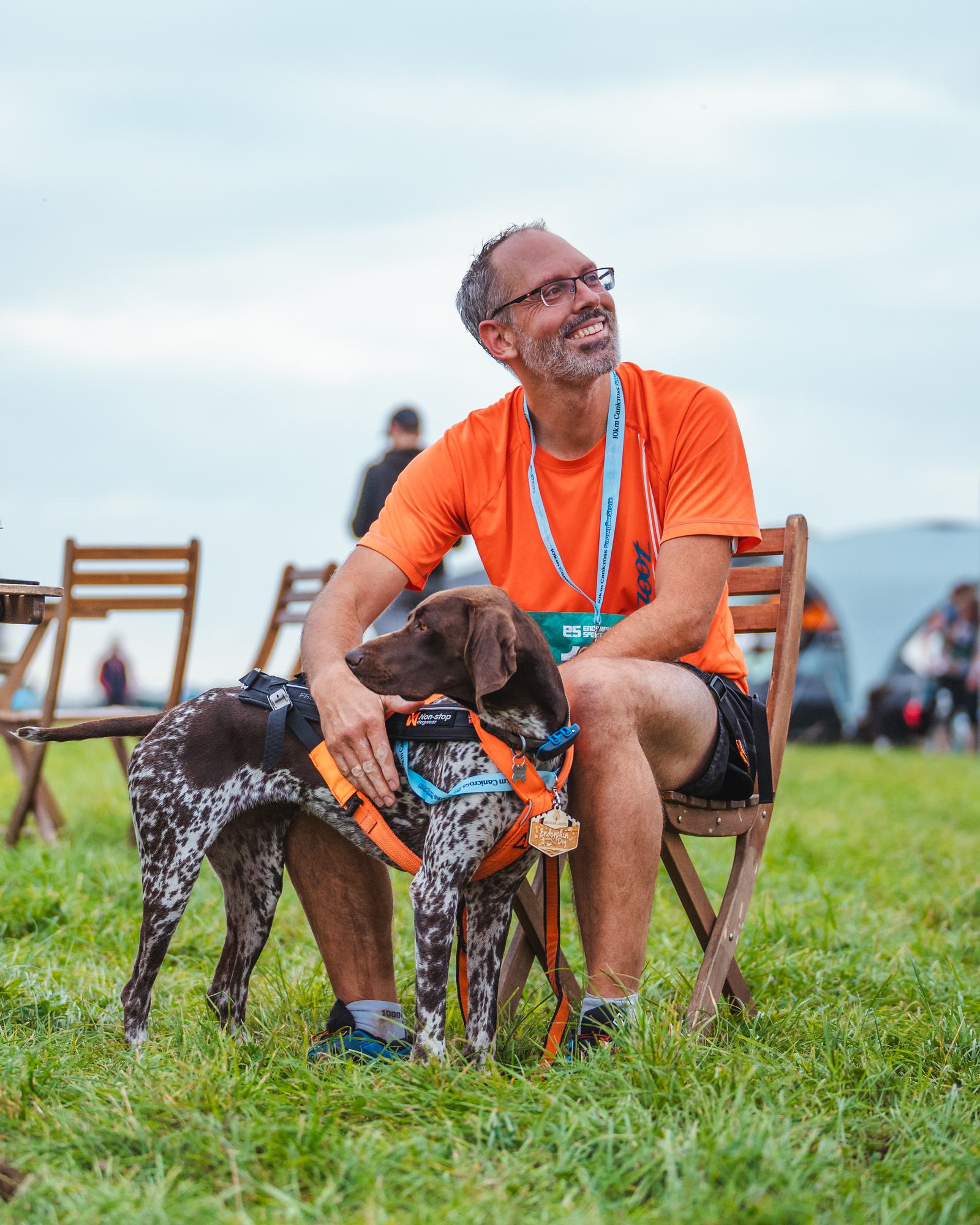 A man in an orange shirt sits on a wooden chair in a grassy field, smiling while petting a brown and white dog wearing an orange vest. The sky is overcast, and other chairs and people are in the background.