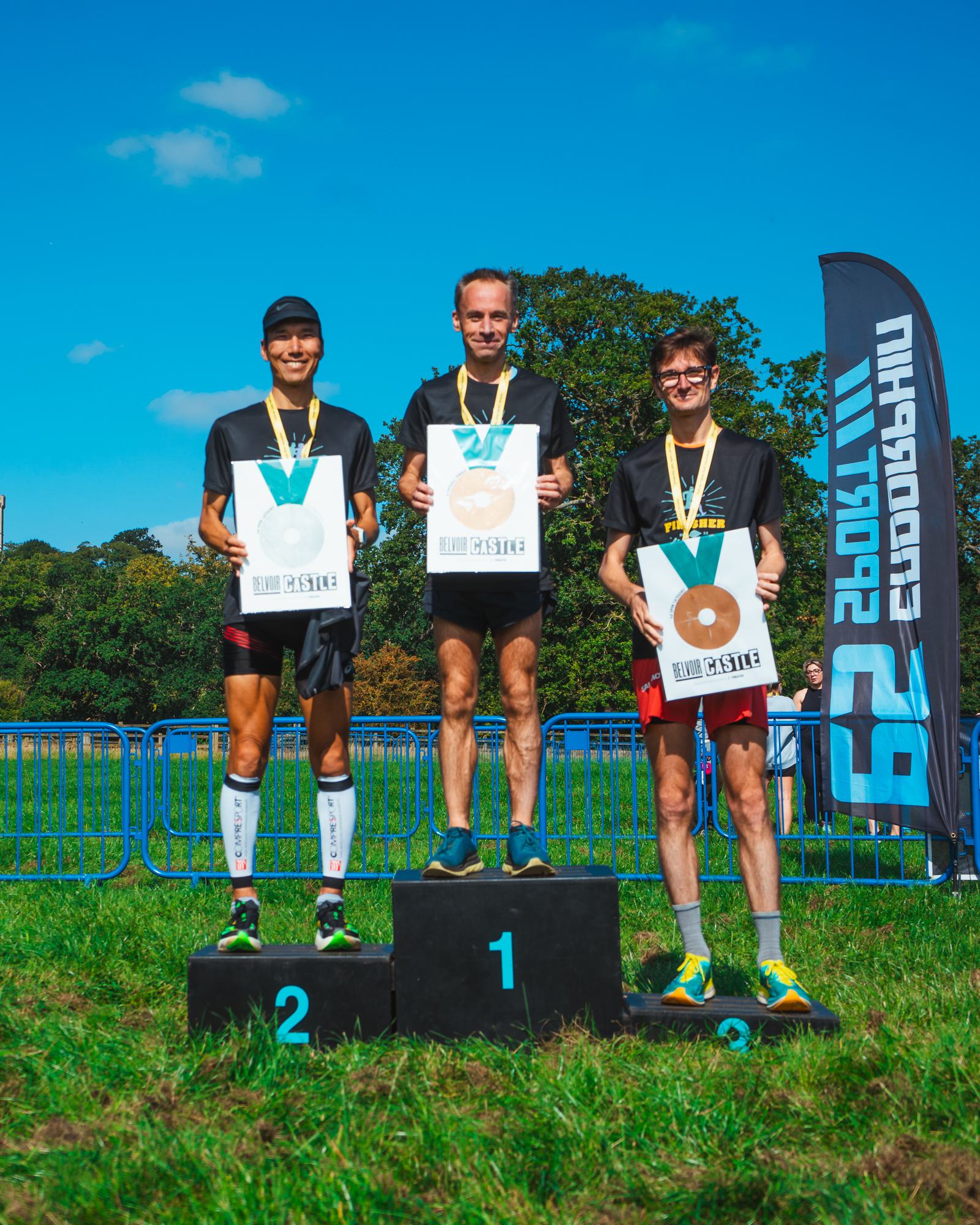 Three runners stand on a podium with medals and certificates in front of a blue sky. The first-place winner is in the center on the highest platform, flanked by the second and third-place finishers. A banner is visible in the background.