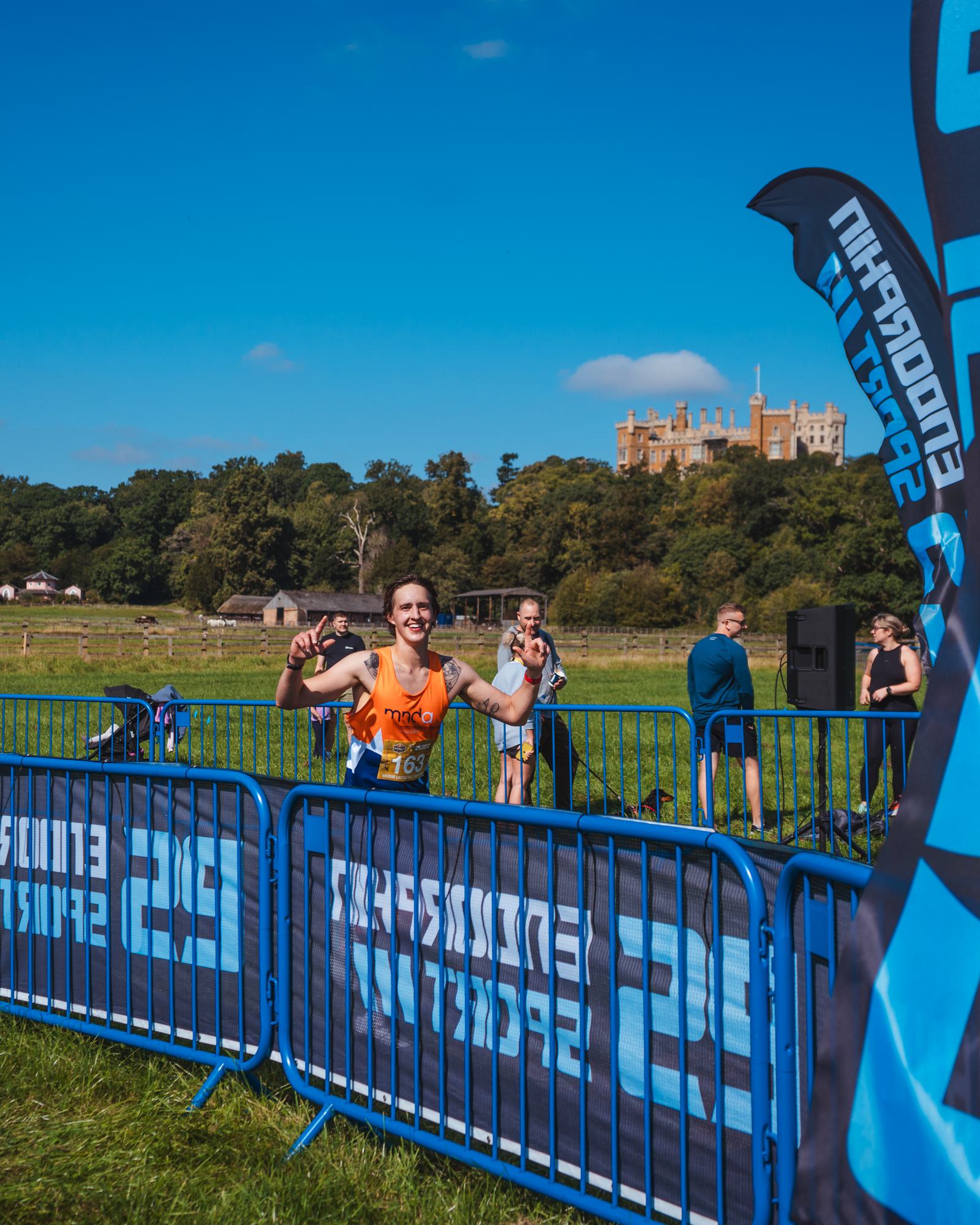 A person wearing a running outfit approaches a finish line with blue banners. They are making a thumbs-up gesture and smiling. In the background, there is a grassy field with trees and a large building. The sky is clear and blue.