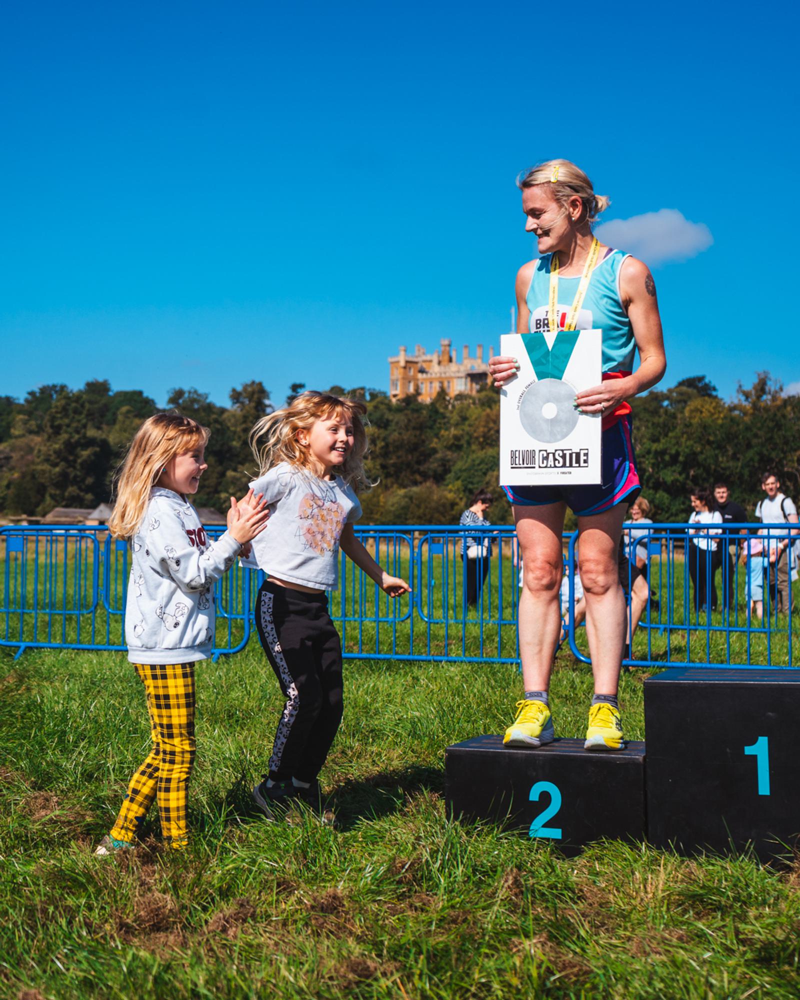 A runner stands on a podium in second place, holding a silver award. Two smiling children stand nearby, celebrating with her. A large, castle-like building is visible in the background under a clear blue sky.