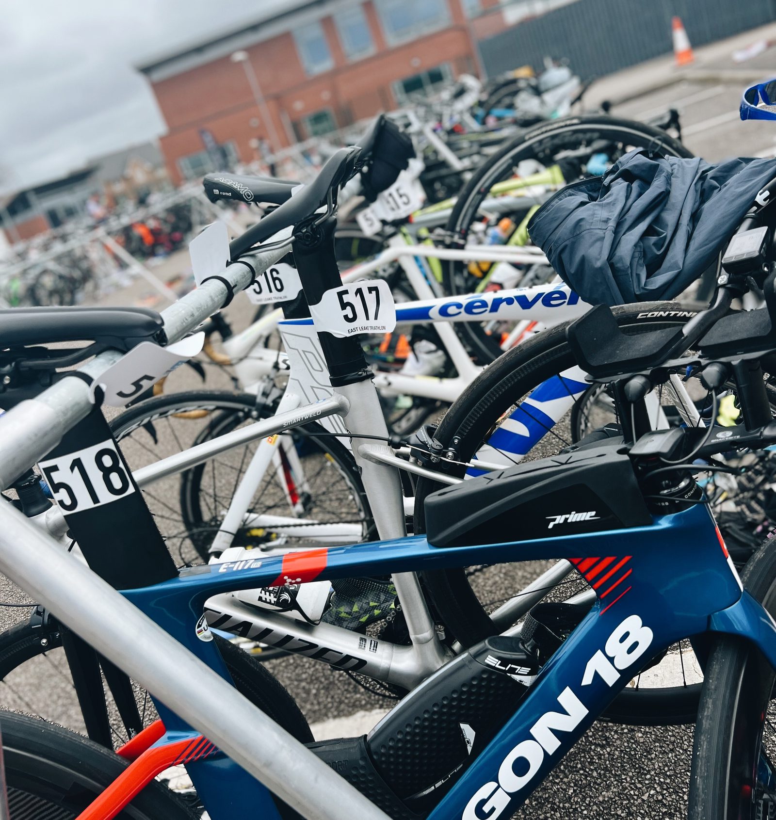 A group of numbered bicycles, including models from brands like Cervélo and Argon 18, are parked and secured in a bike rack outside. A brick building is visible in the background. Some of the bikes have gear such as jackets or bags attached to them.