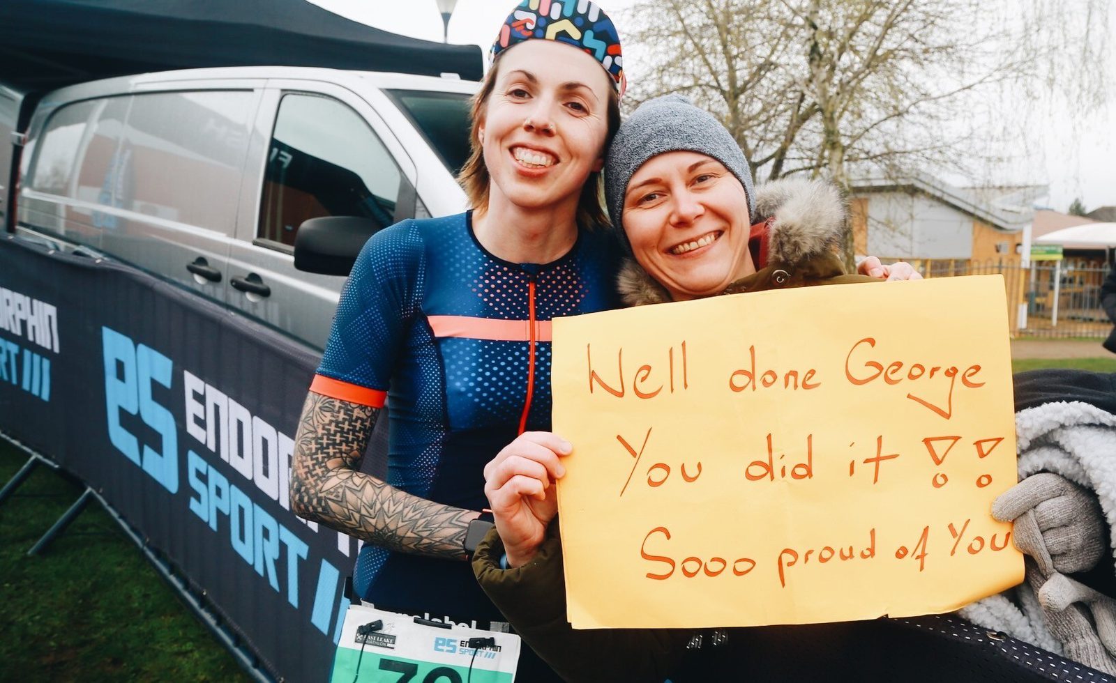 Two people smiling and posing outdoors. The person on the left is in athletic gear, while the person on the right holds a bright yellow sign that reads, "Well done George, You did it! Sooo proud of you!" They stand in front of a van with "ENDURAN SPORT" branding.