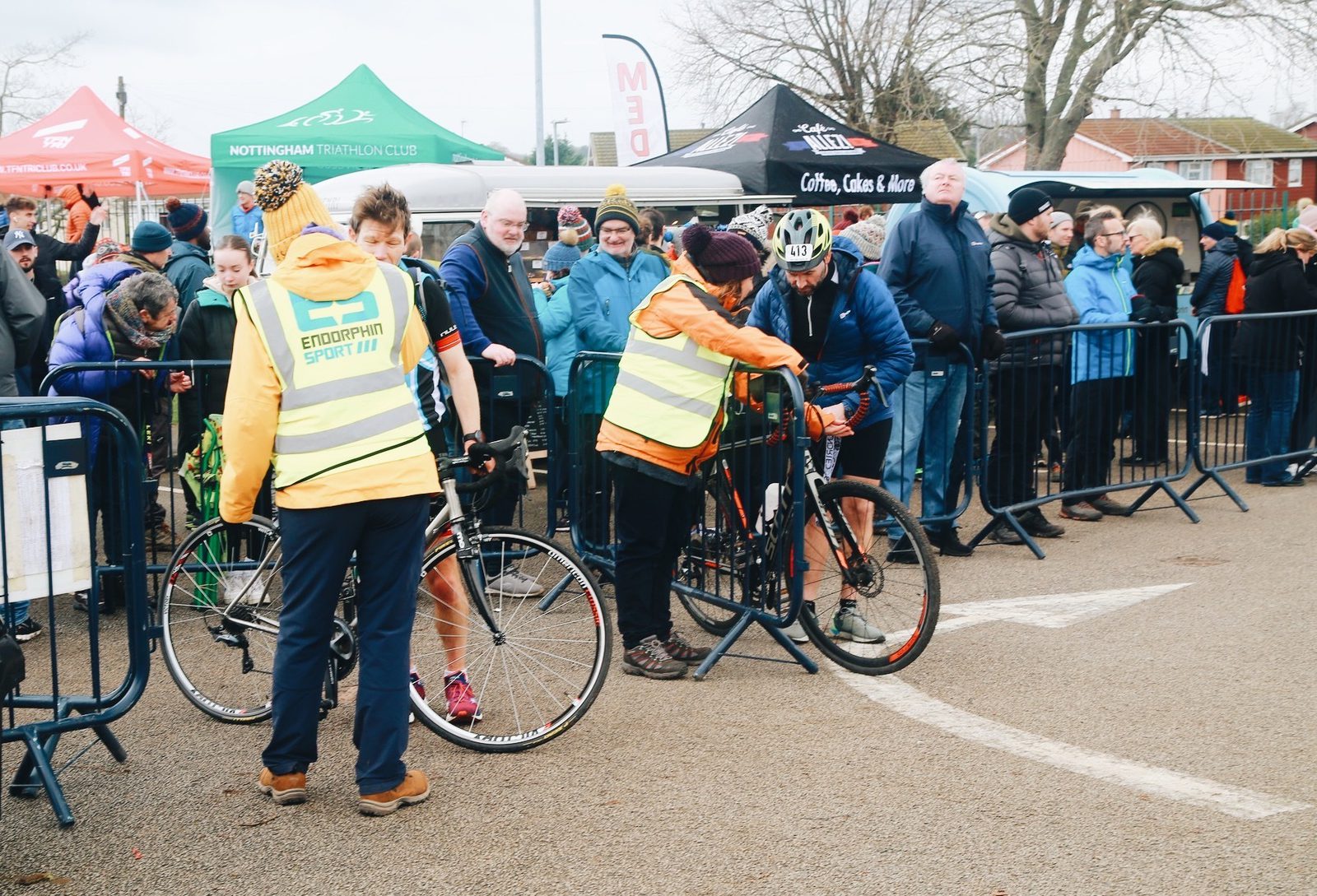 A group of cyclists at a race event are interacting with volunteers dressed in high-visibility vests. Barriers are set up along the track, and spectators are gathered behind them. Tents and banners from various clubs and vendors are visible in the background.