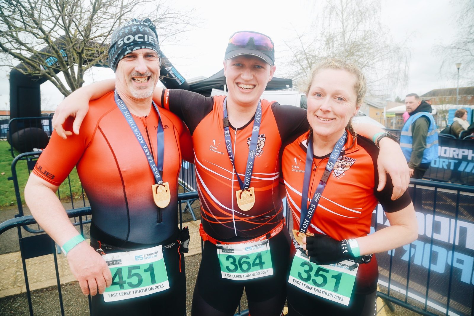 Three triathletes stand in a row with their arms around each other, smiling broadly. They each wear a medal around their necks, race bibs numbered 451, 364, and 351, and sport matching red and black gear. A tree and other participants are visible in the background.